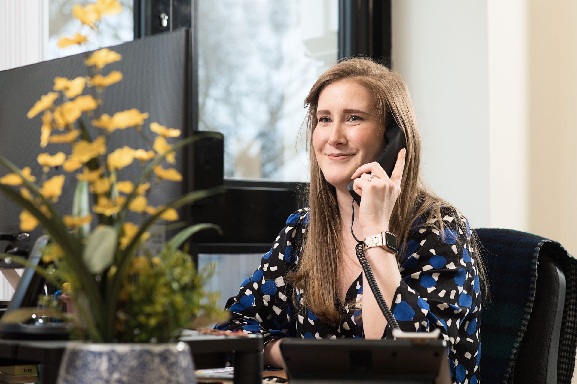A woman at work talks on a corded telephone, smiling warmly. A computer monitor and a blurry potted orchid plant with yellow flowers are visible on her desk in the foreground.