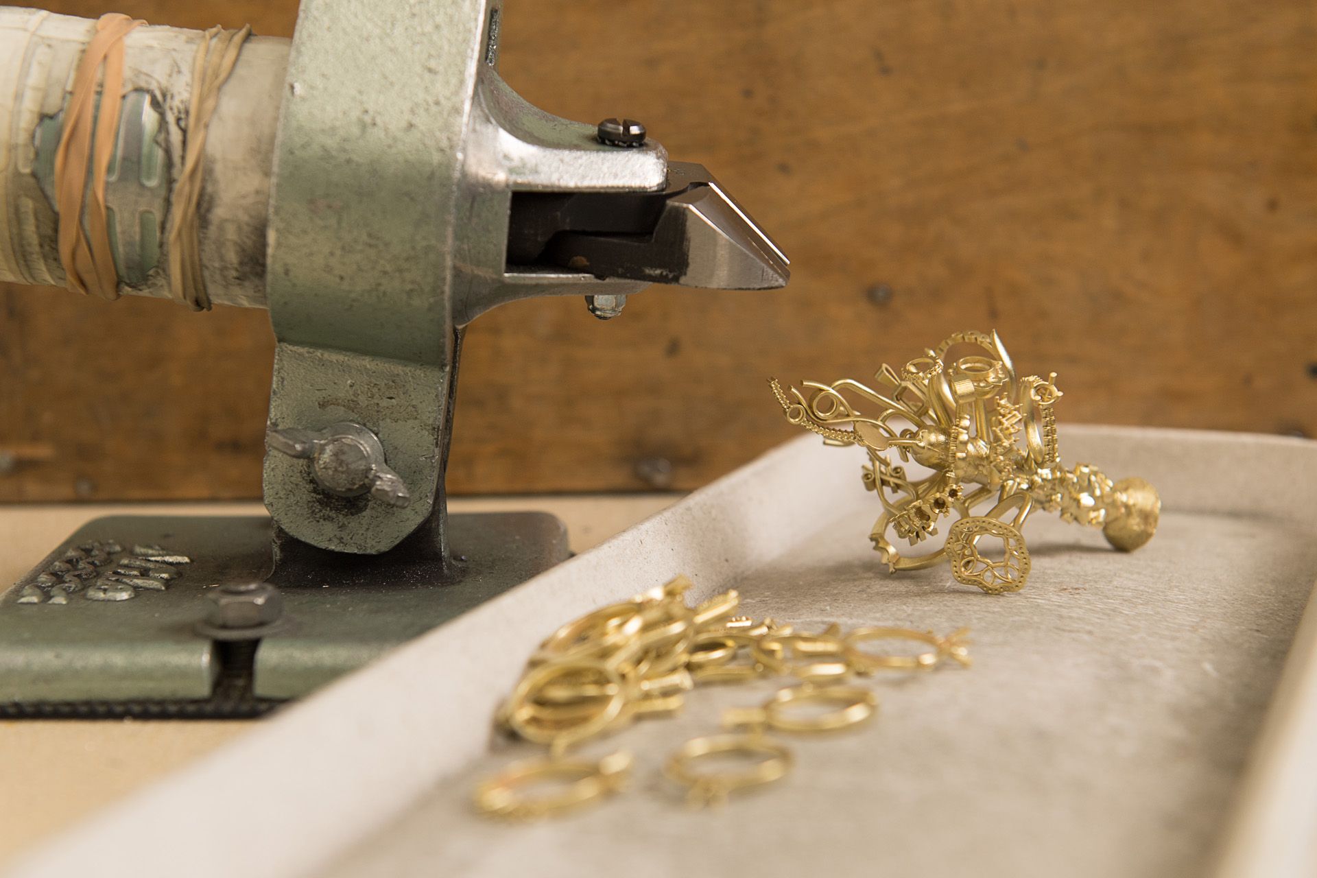 A close-up shot of a goldsmith's workbench showing several unfinished gold rings and a small, intricate gold piece next to a piece of metalworking equipment.