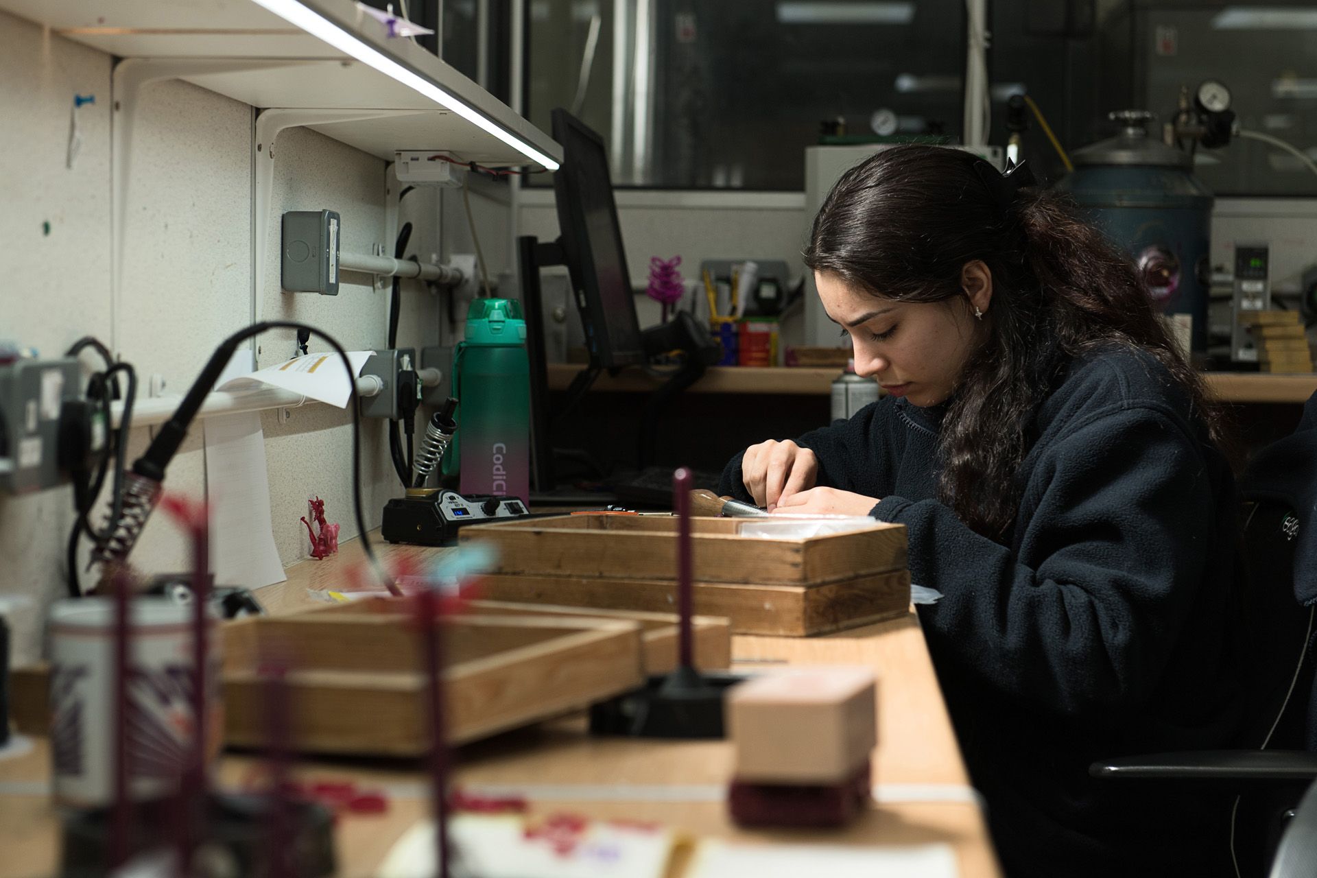 A jeweller with long, dark hair is focused on their work at a busy workbench, with various tools and equipment surrounding them.