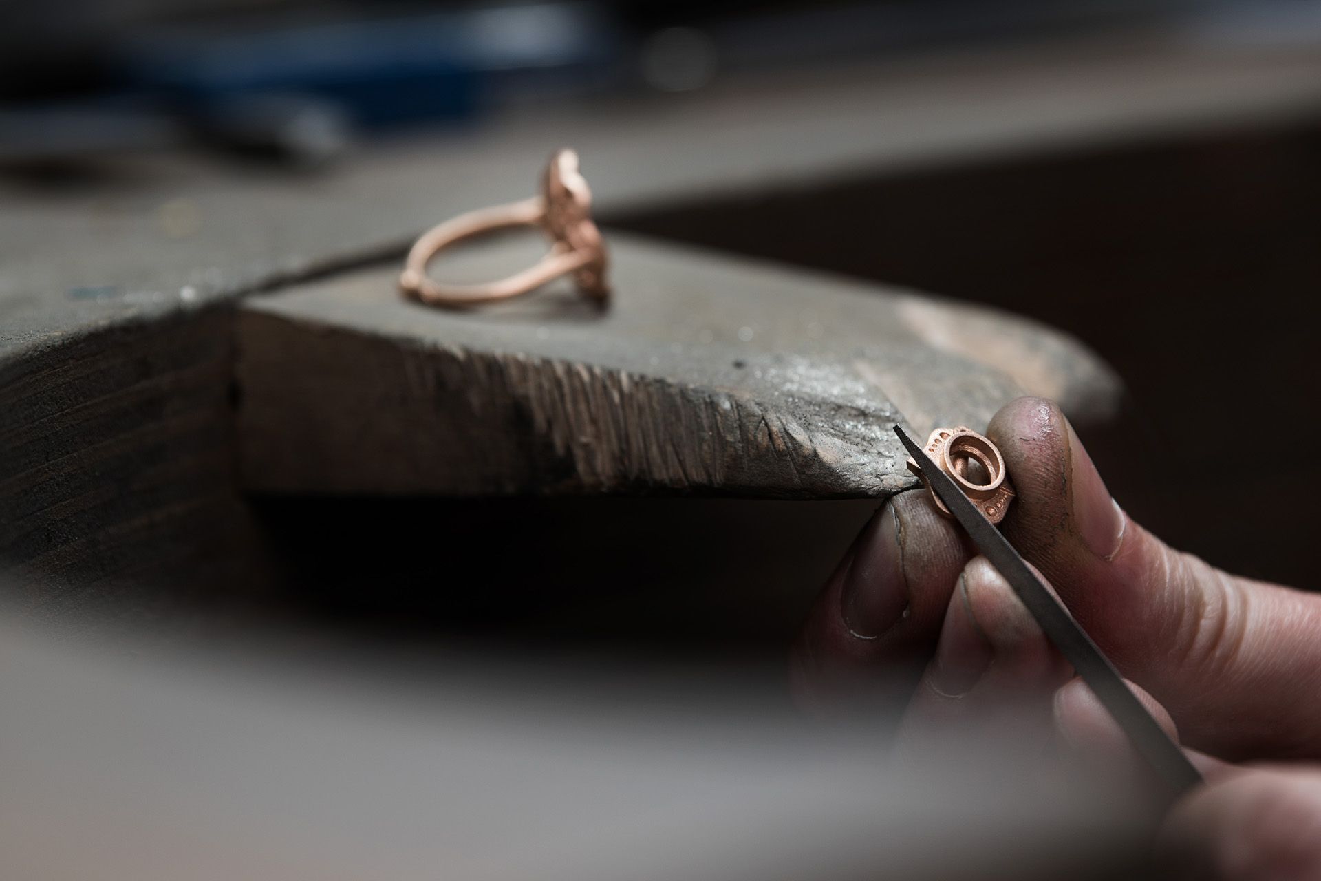 A jeweller's hand uses tweezers to carefully hold and position a small piece of jewellery at a workbench. In the background, a finished ring sits on a bench pin.