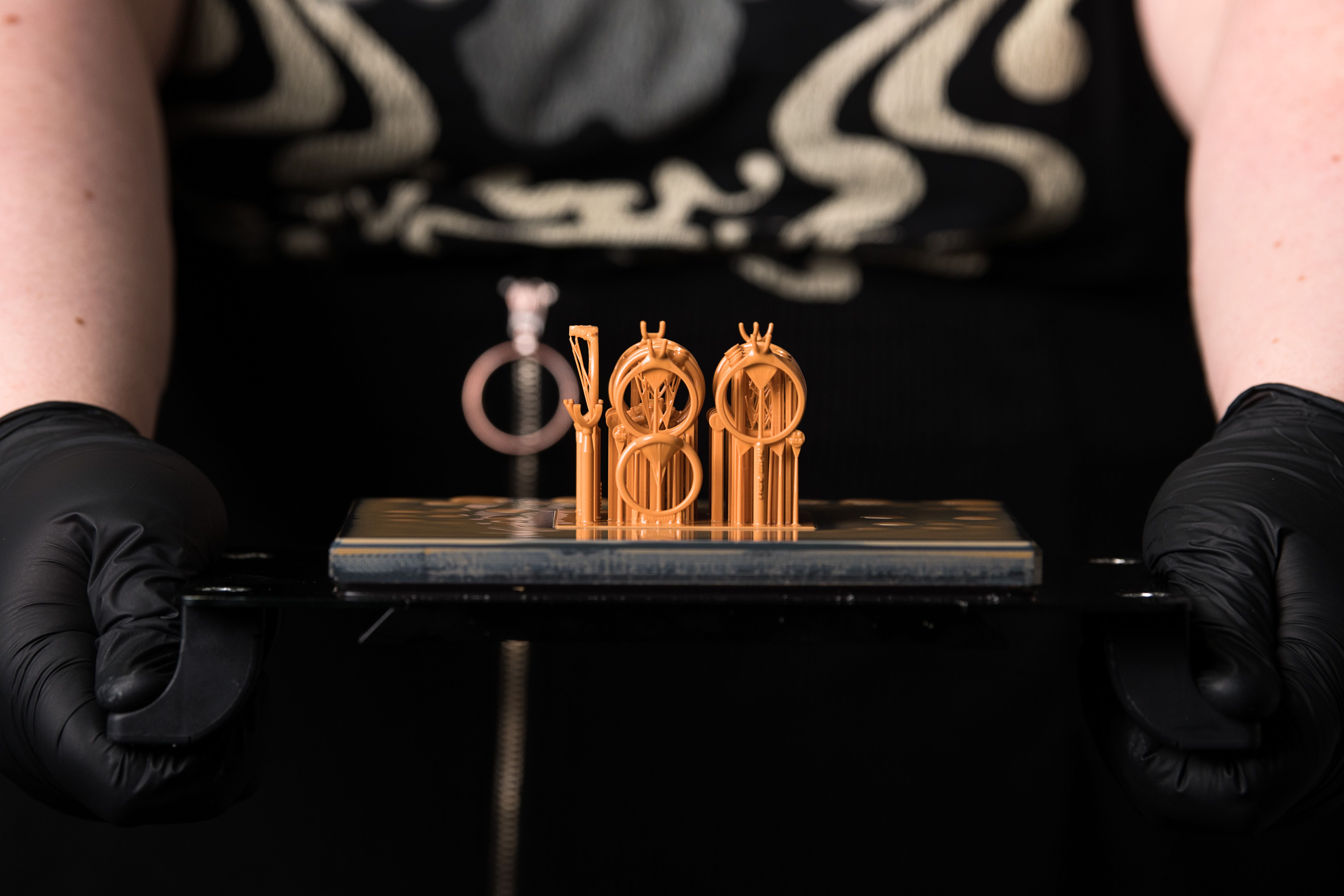 Person wearing black gloves holds a tray with 3D-printed ring designs in orange resin. Background features black clothing with white patterns.