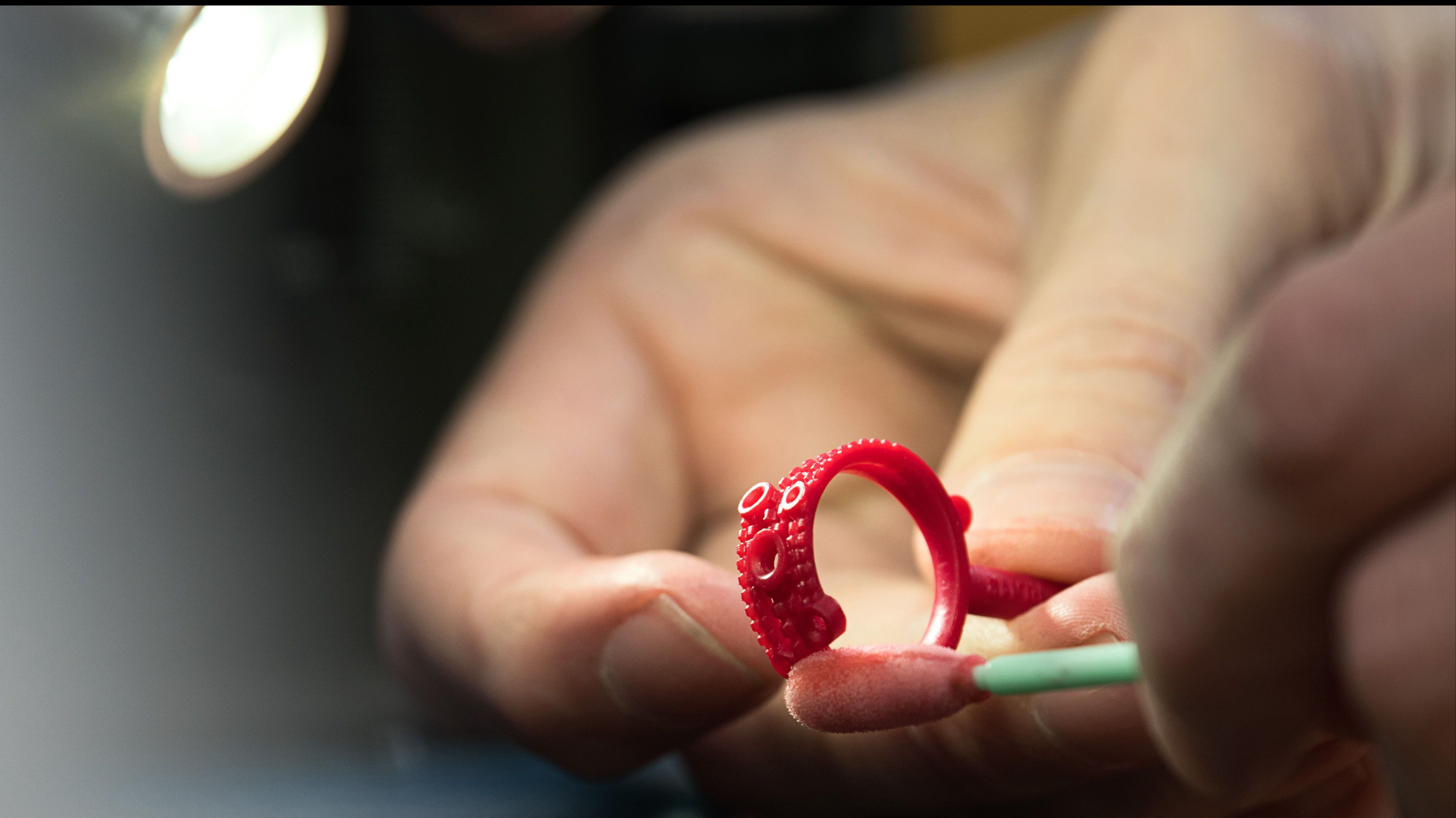 A close-up shot of a jeweller’s hands holding a vibrant red wax model of a ring, carefully cleaning it with a cotton bud.