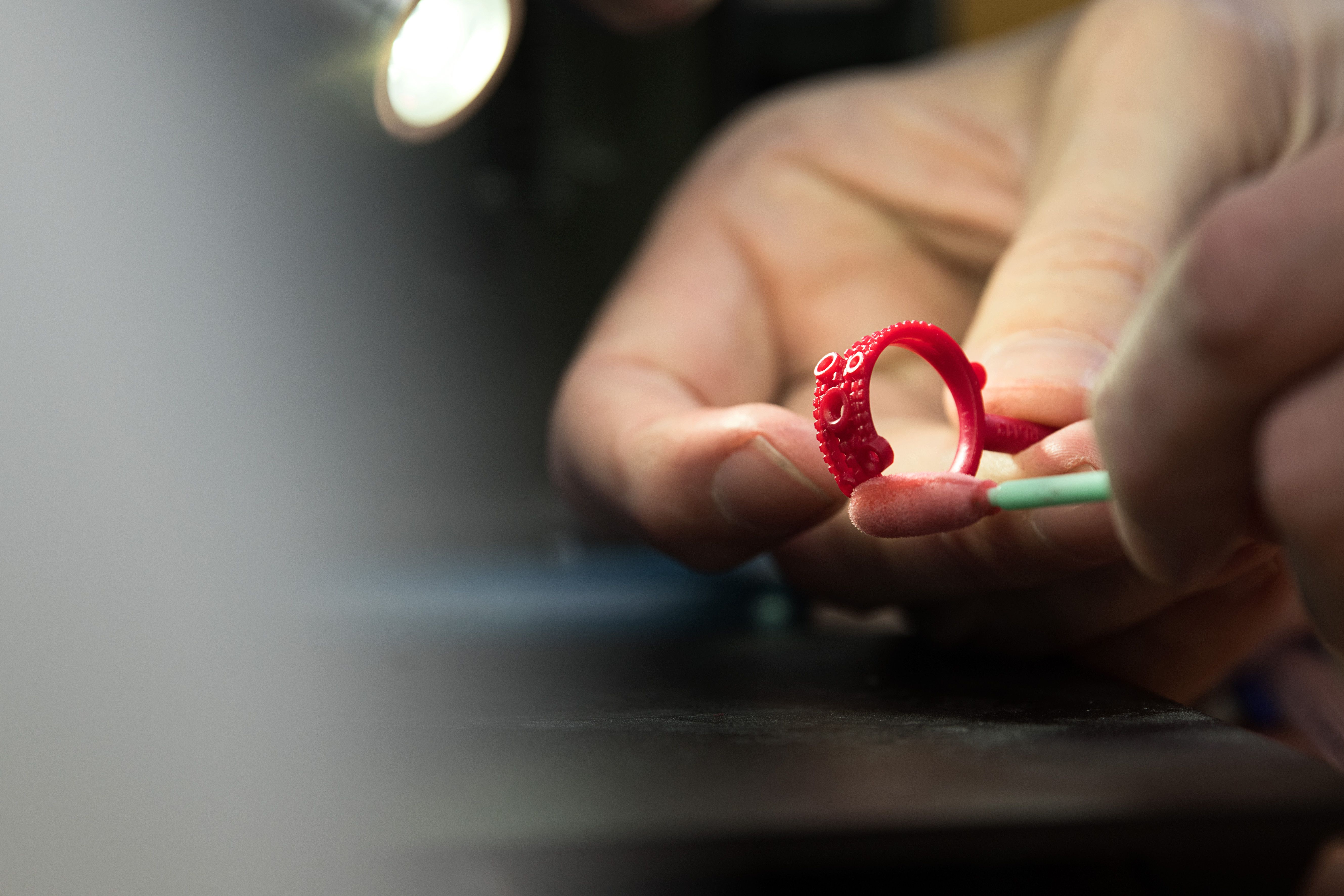 A close-up shot of a person's hands holding a red wax model of a ring with a detailed, textured surface. They are using a small, light-colored tool to work on the ring under the focused beam of a light.
