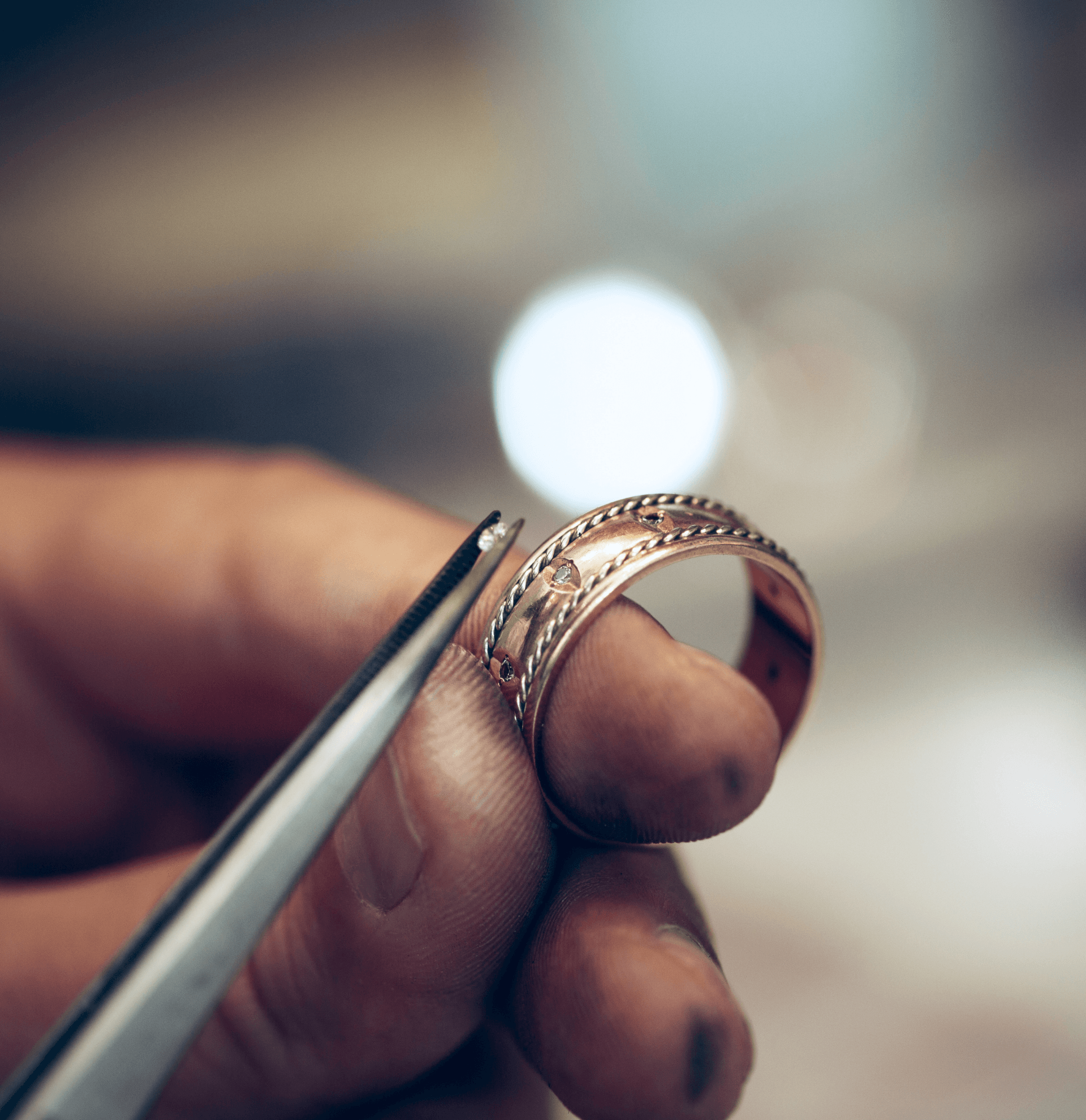Close-up of a jeweler’s hand holding an ornate gold or rose gold ring, while using tweezers to adjust a small detail or gemstone.