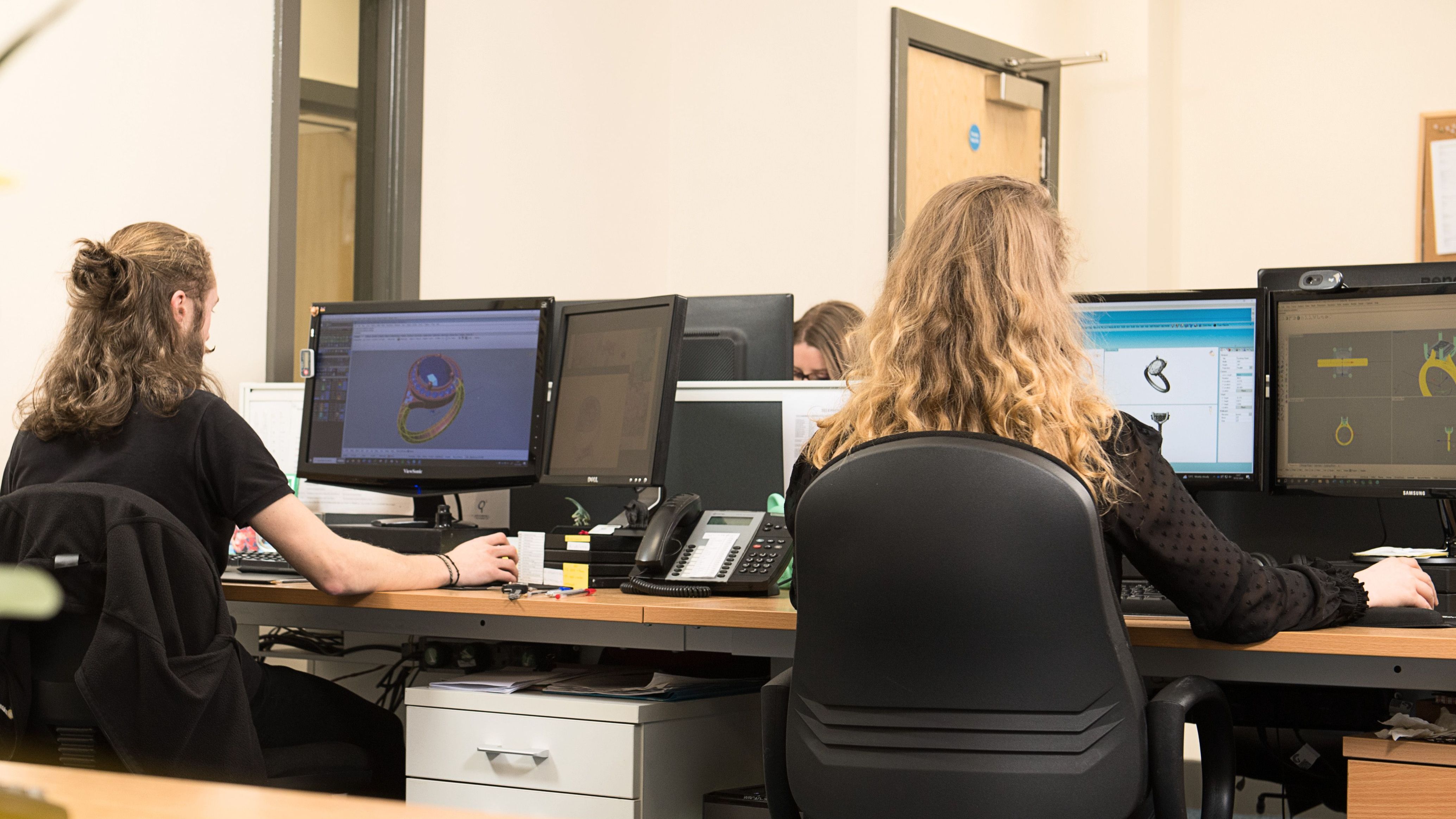 A view from behind two people sitting at a long desk in an office. They are both working on multiple computer monitors