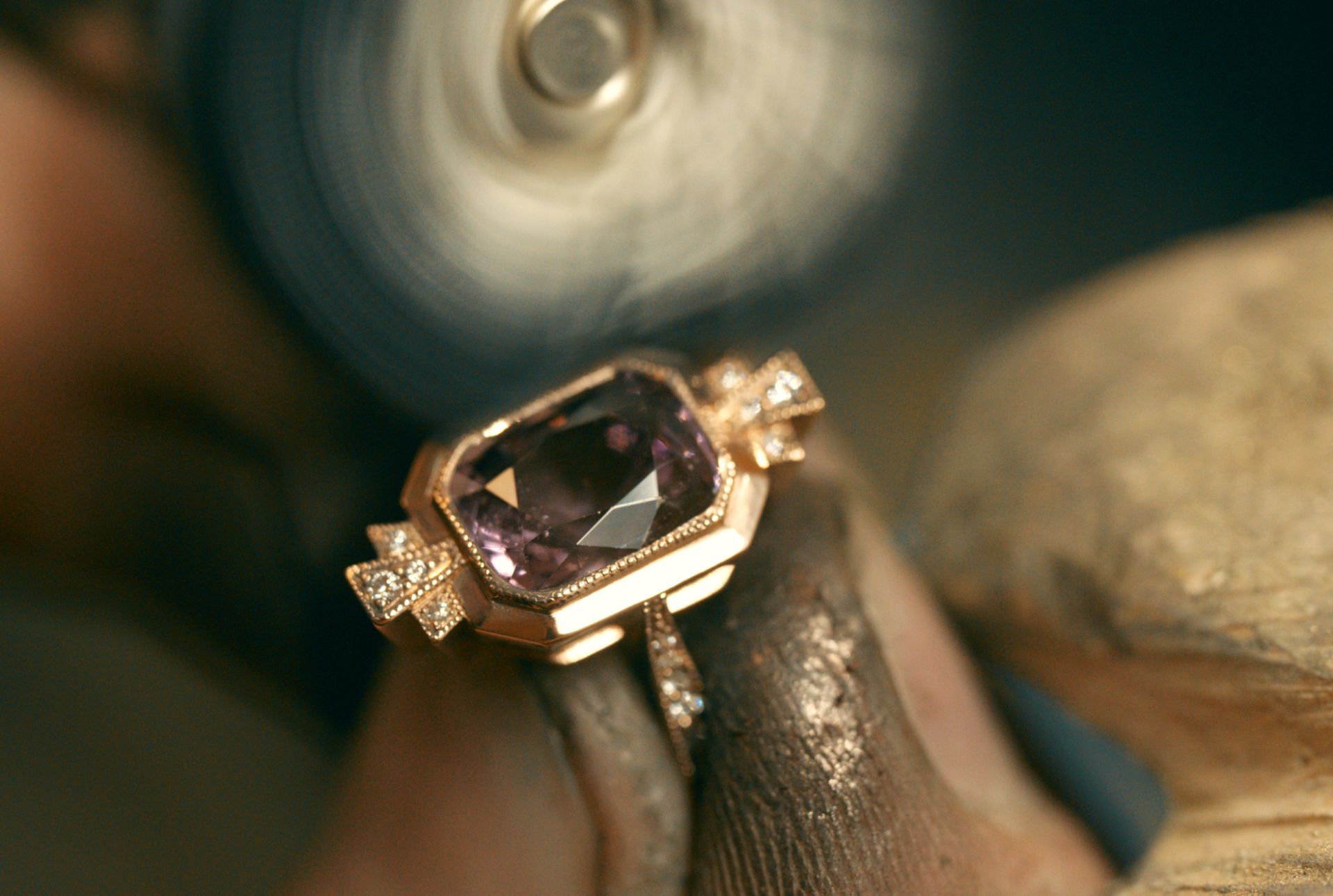 A close-up shot of a ring with a large, faceted purple gemstone and small accent diamonds being held against a spinning polishing wheel.