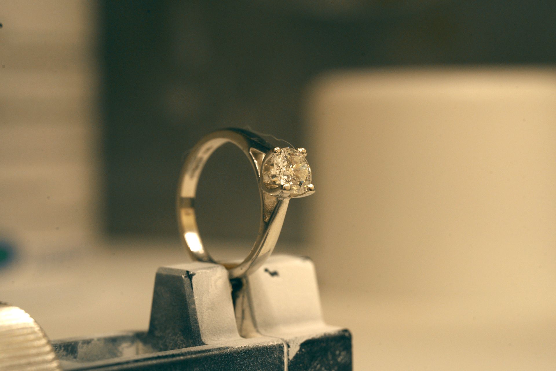 A silver ring with a single square-cut diamond is held upright by a small clamp. The background is a blurry workshop or studio.