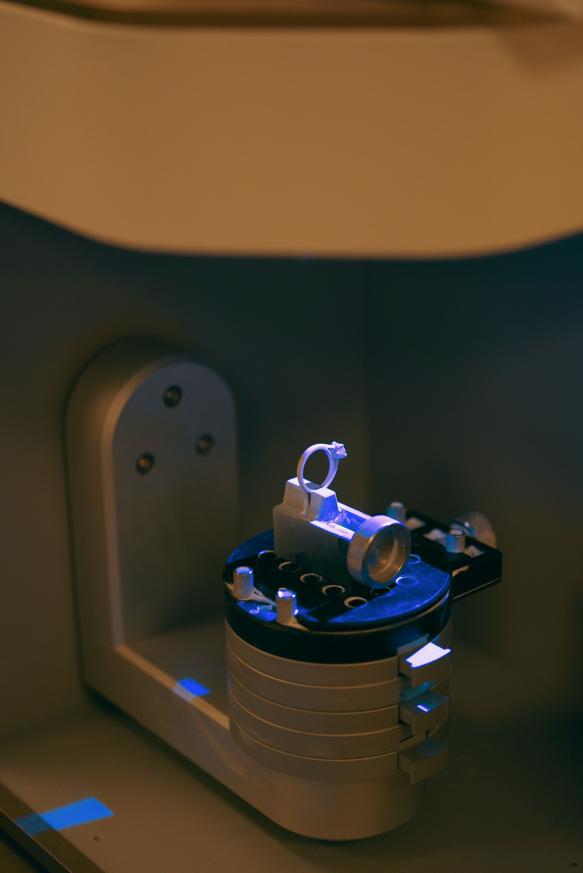 A detailed ring design is visible, held in place by the machine's chuck, with bright blue light from the machine shining on it.