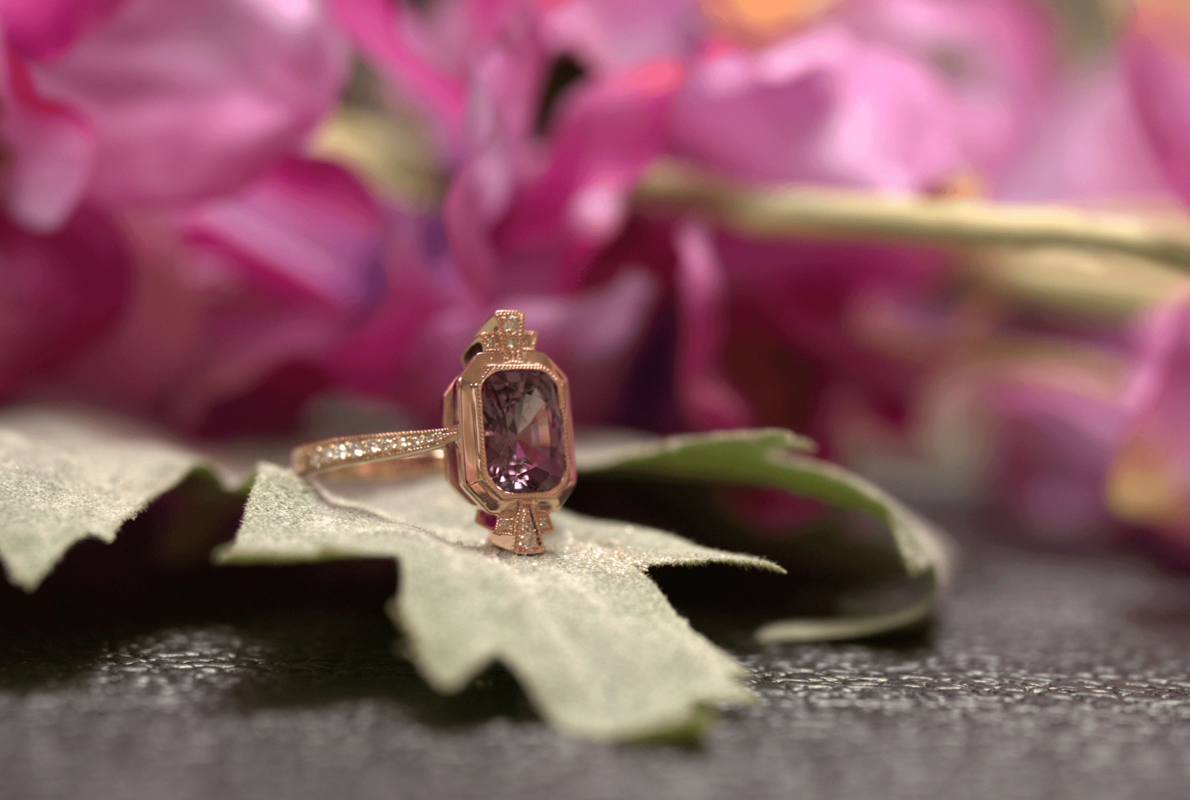 A rose gold ring with a purple gemstone rests on a green leaf, set against a soft-focus pink floral background, evoking elegance and romance.