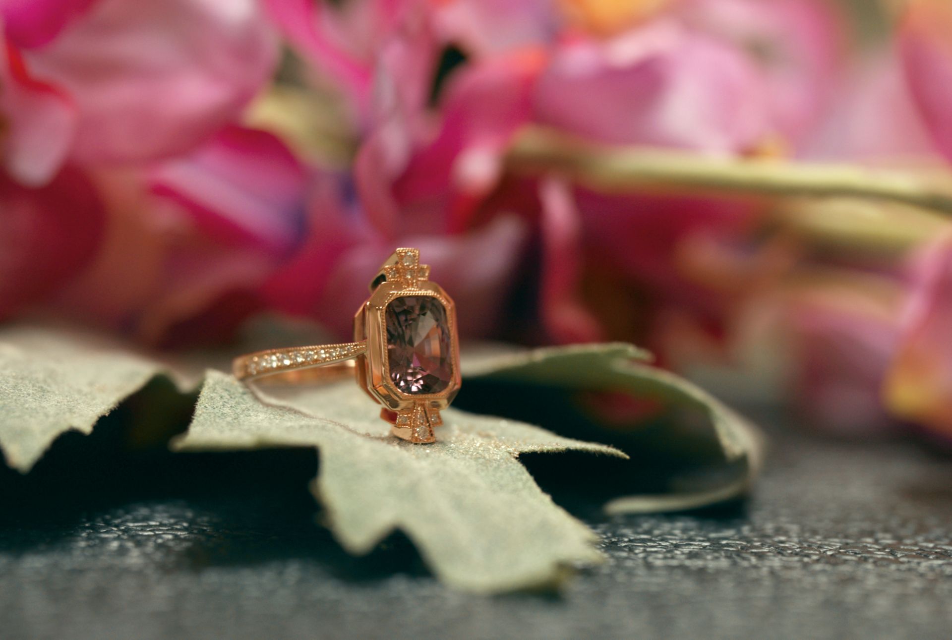 A rose gold ring with a purple gemstone rests on a green leaf, set against a soft-focus pink floral background, evoking elegance and romance.