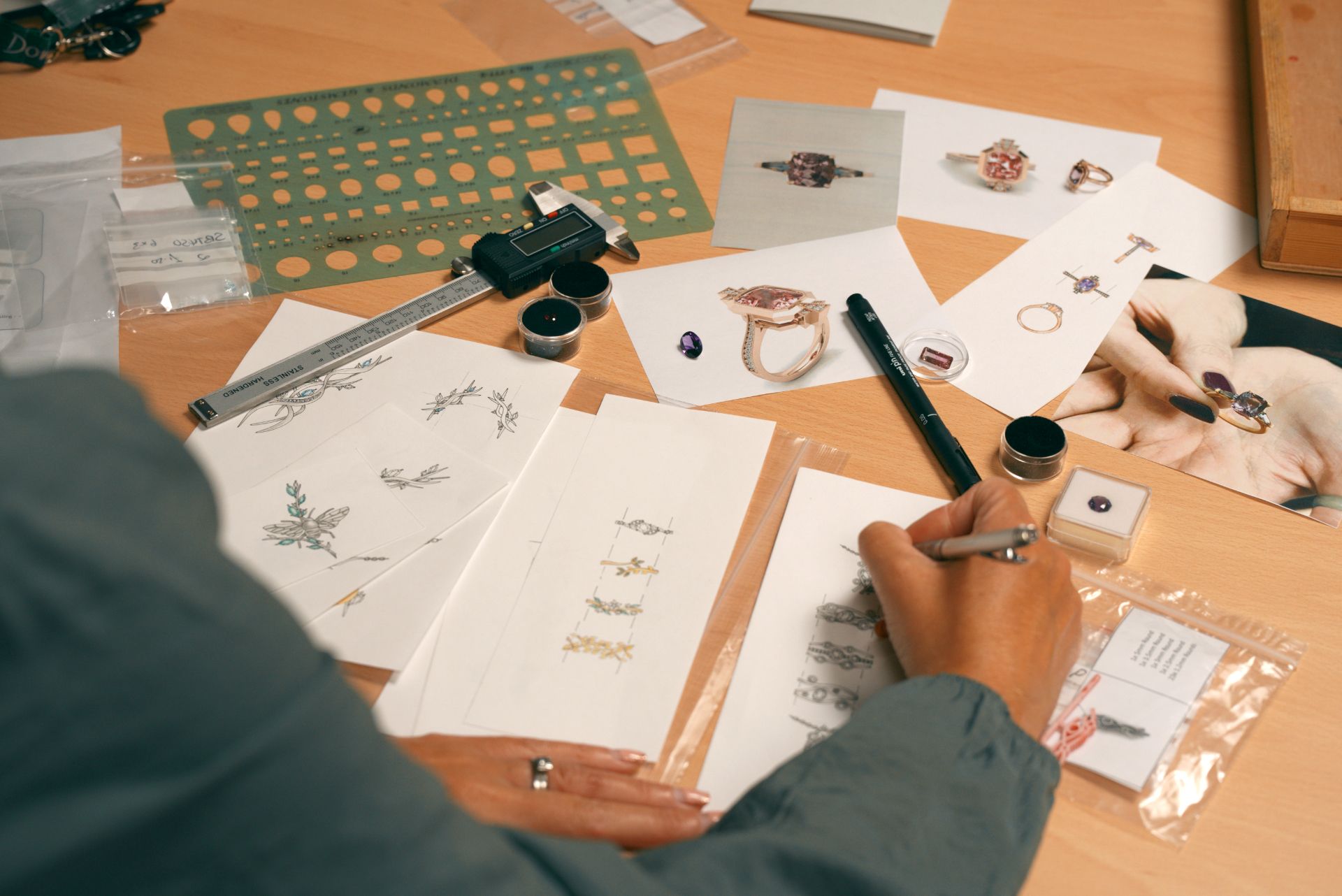 An artist sketches jewelry designs on paper, surrounded by tools and photos of rings. The workspace conveys creativity and focus.