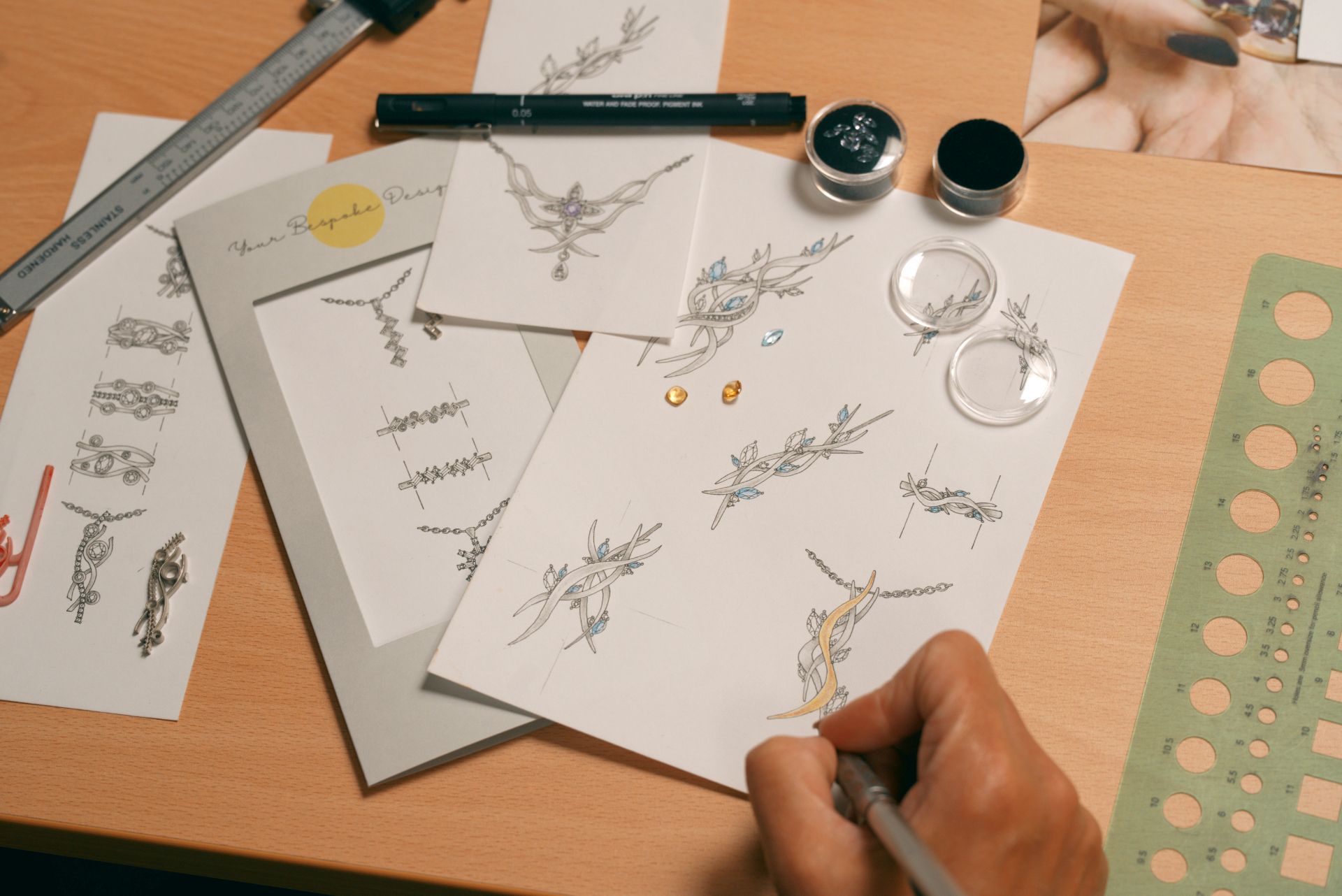 Close-up of a person drawing detailed jewelry concepts, including necklaces and earrings, with various sketches, a ruler, and magnifying tools spread across a workspace.