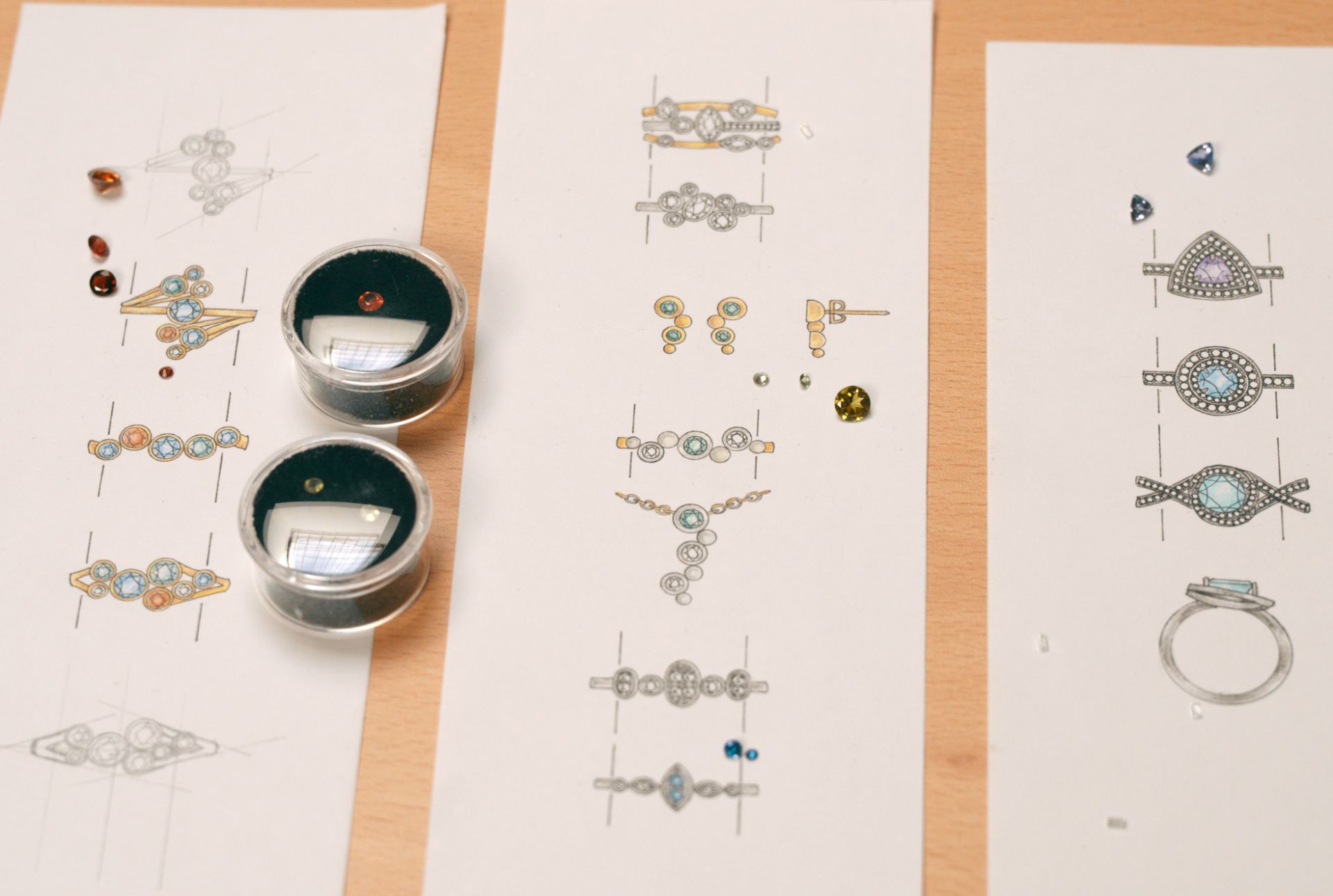 An overhead view of hand-drawn jewellery sketches with small, loose gemstones arranged next to them.