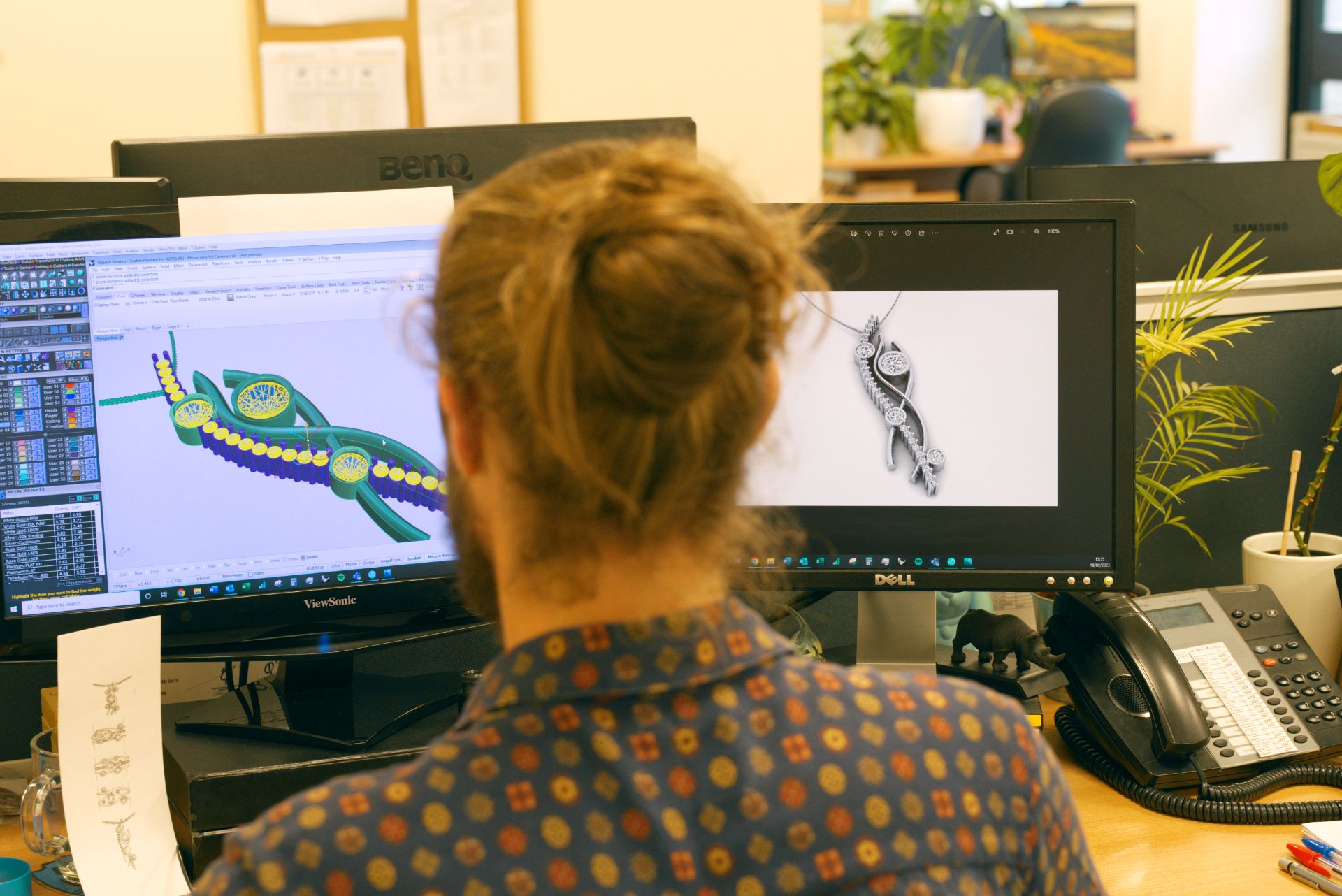 A person sits at a desk, looking at two computer monitors. The left screen shows a colourful, intricate piece of jewellery in a CAD program, while the right screen displays a realistic 3D render of a diamond pendant. 