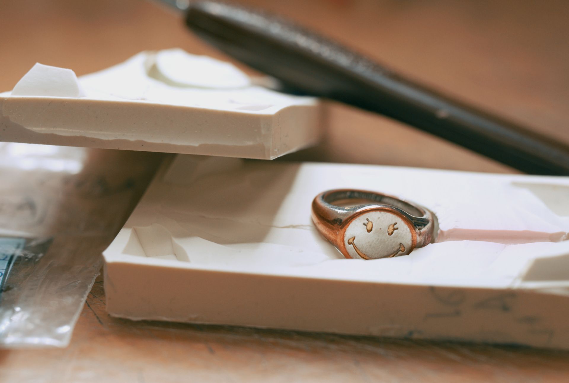 A close-up, horizontal shot of a silver and copper-colored signet ring with a smiley face etched into it, lying on a white, waxy mold. A sculpting tool or craft knife with a dark handle is visible in the background, resting on a wooden surface.