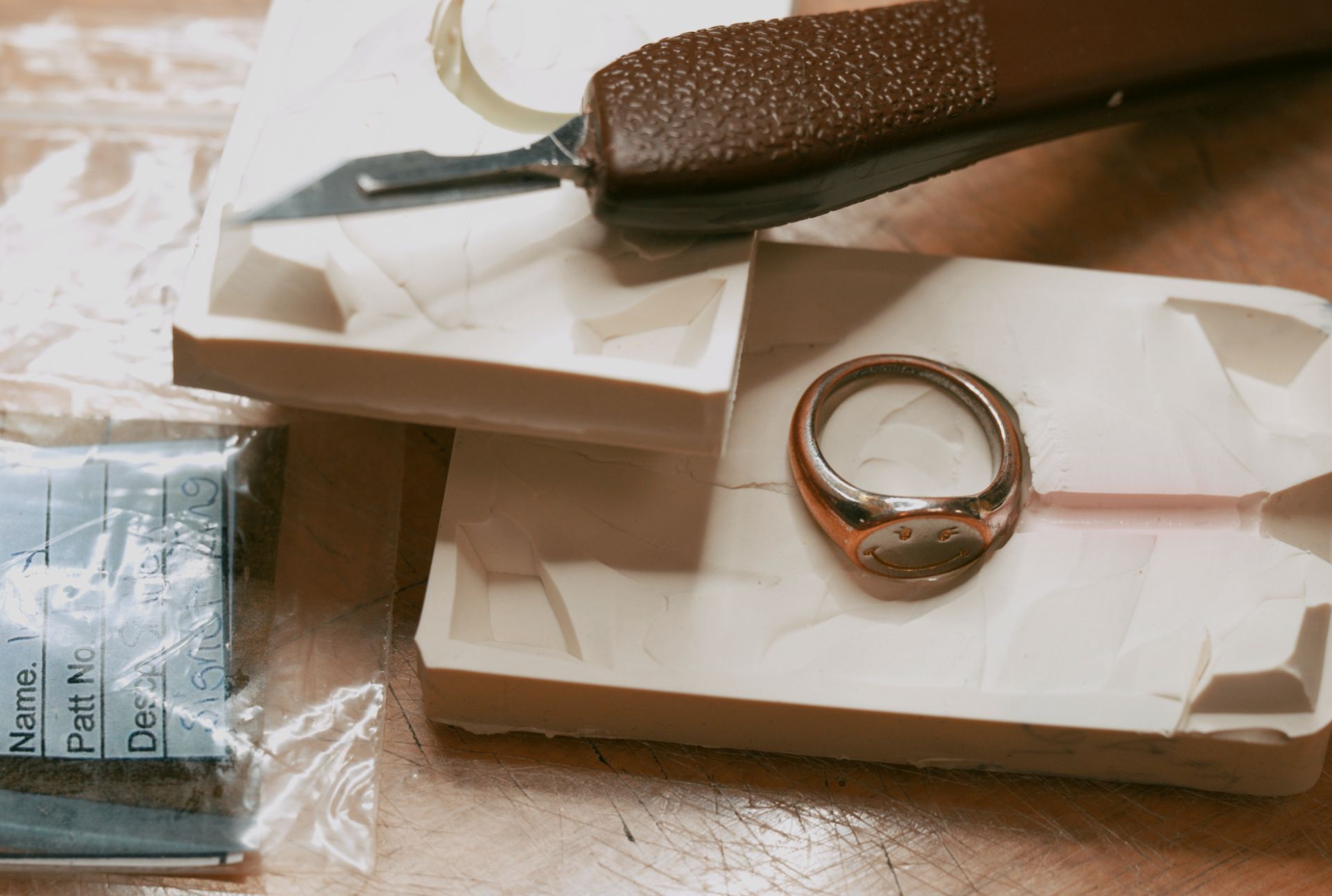 A two-toned smiley face signet ring rests on a white wax mold, with a craft knife and a second mold in the background.