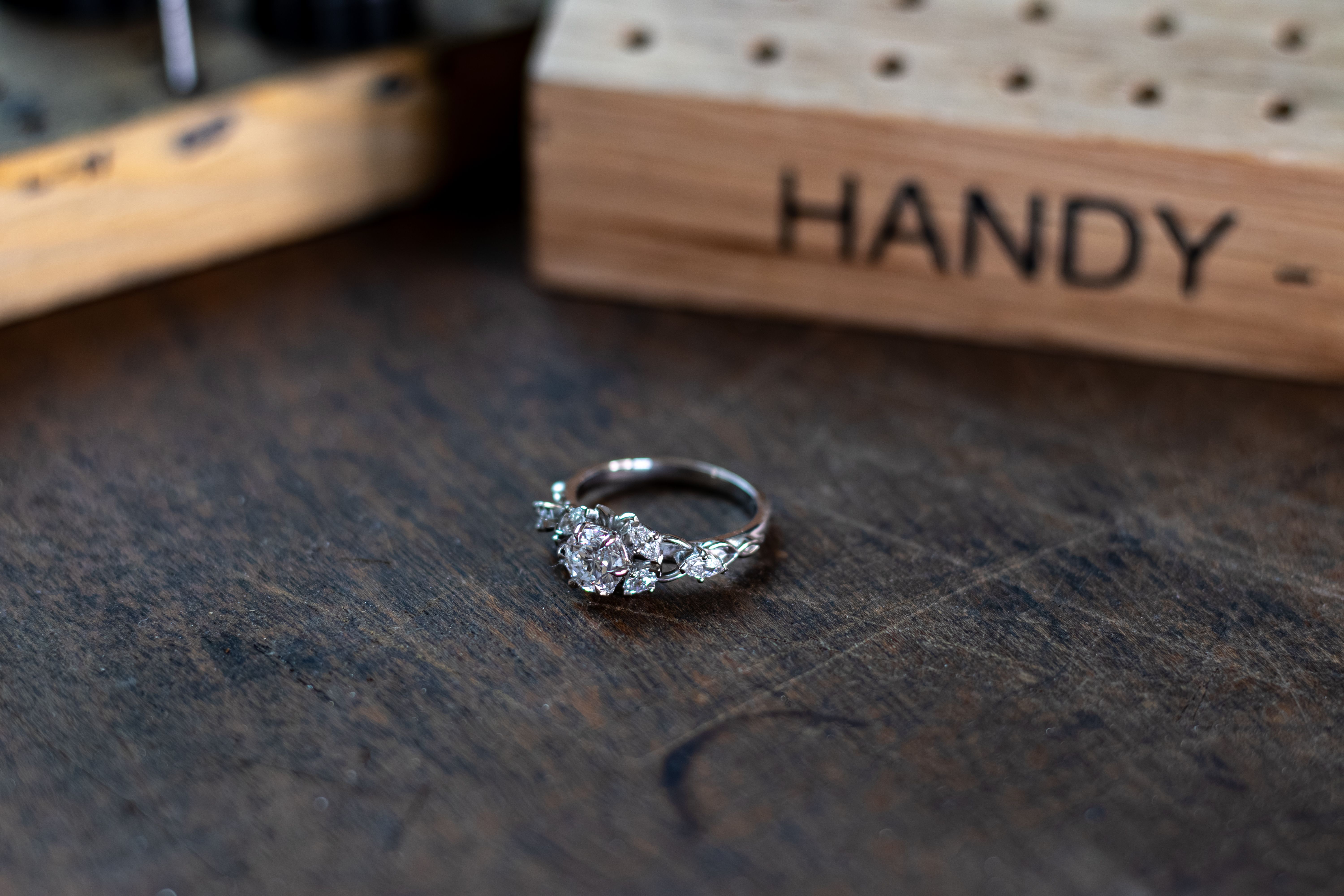 A silver ring with a large pink central gemstone and smaller clear stones set into an intricate floral design rests on a dark wooden surface, with a blurred wooden tool holder in the background.