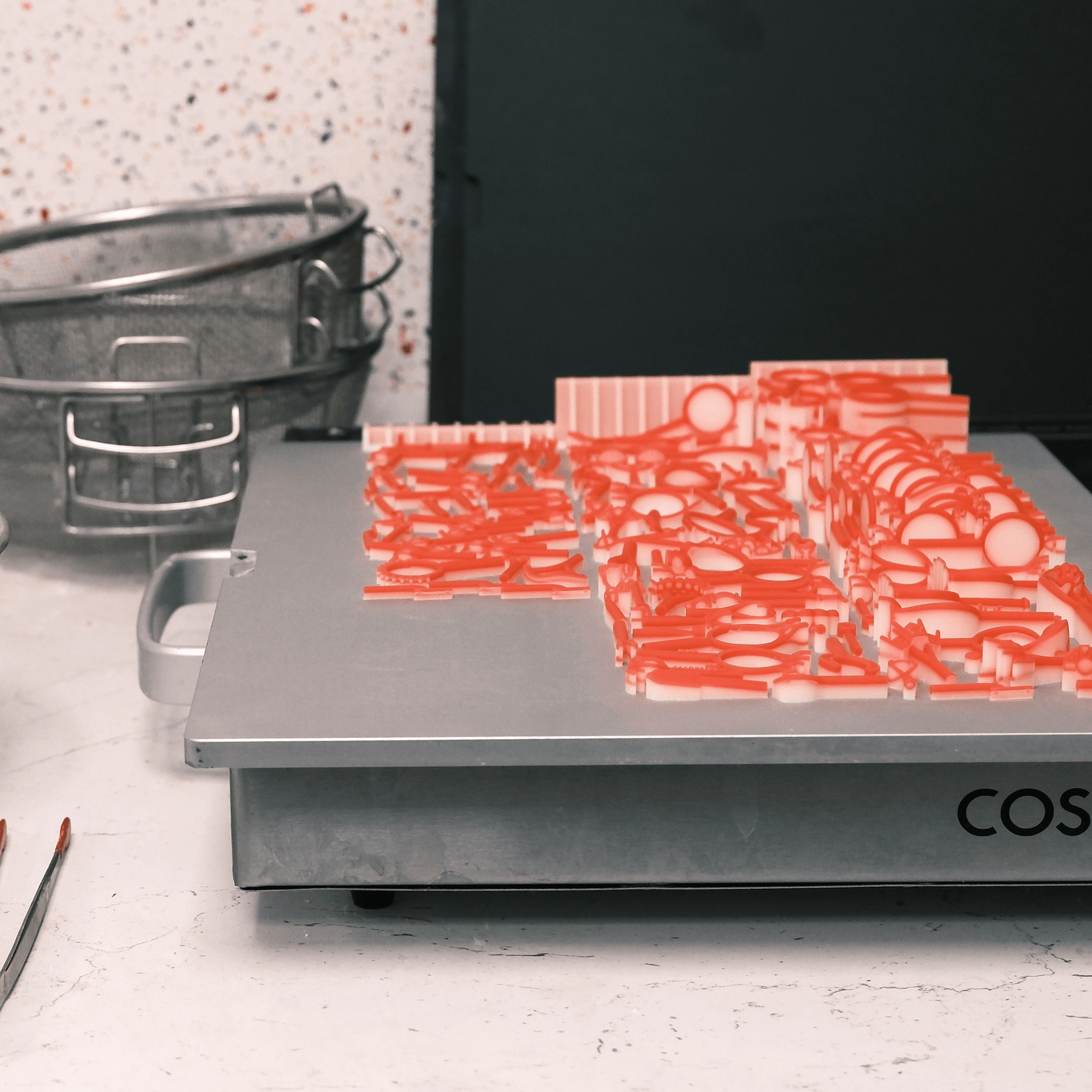 A metal platform covered with numerous red 3D-printed wax or resin jewellery patterns, next to a metal sieve.