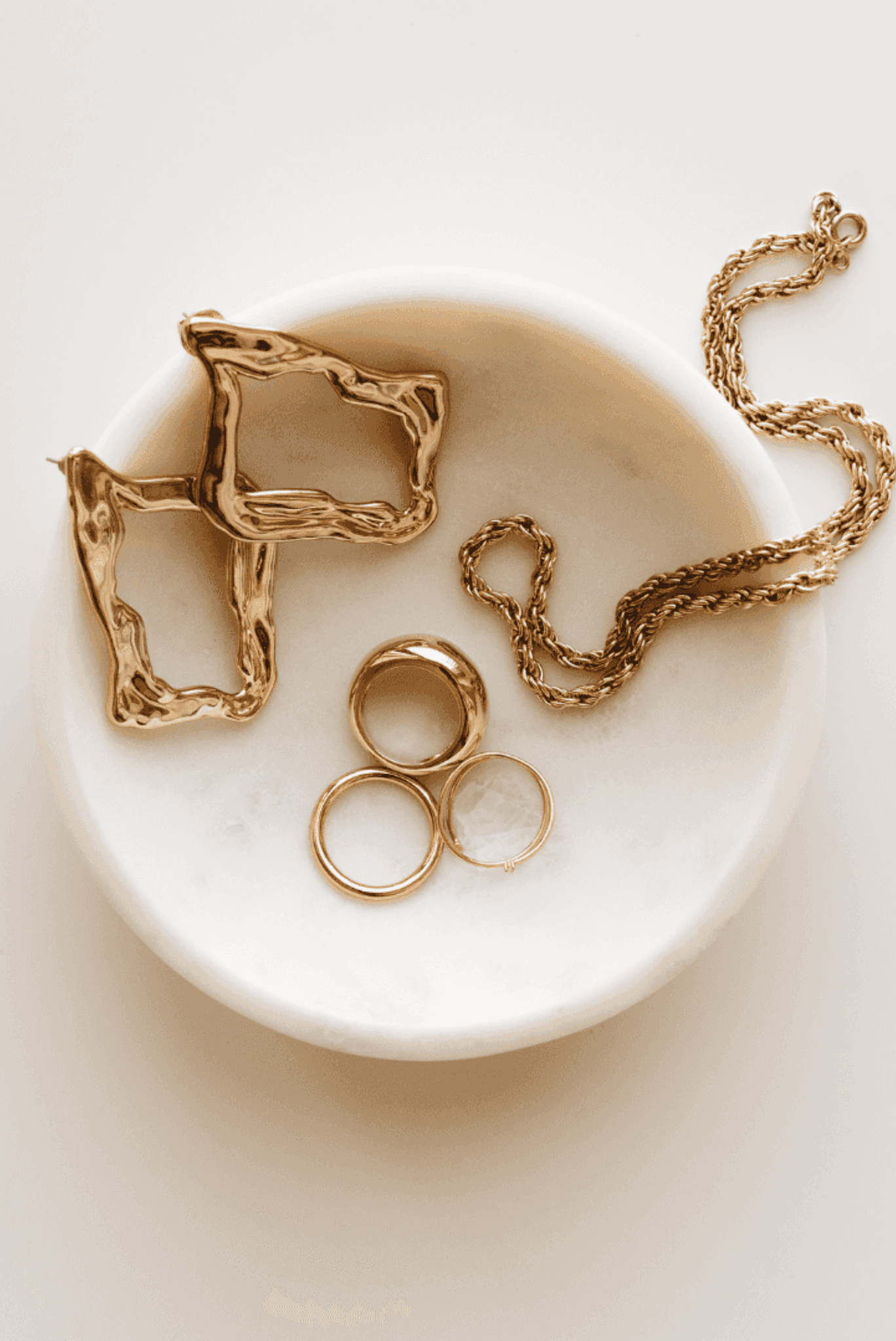Three pieces of jewellery include a pair of chunky, organic-shaped hoop earrings, three different rings, and a rope chain necklace.