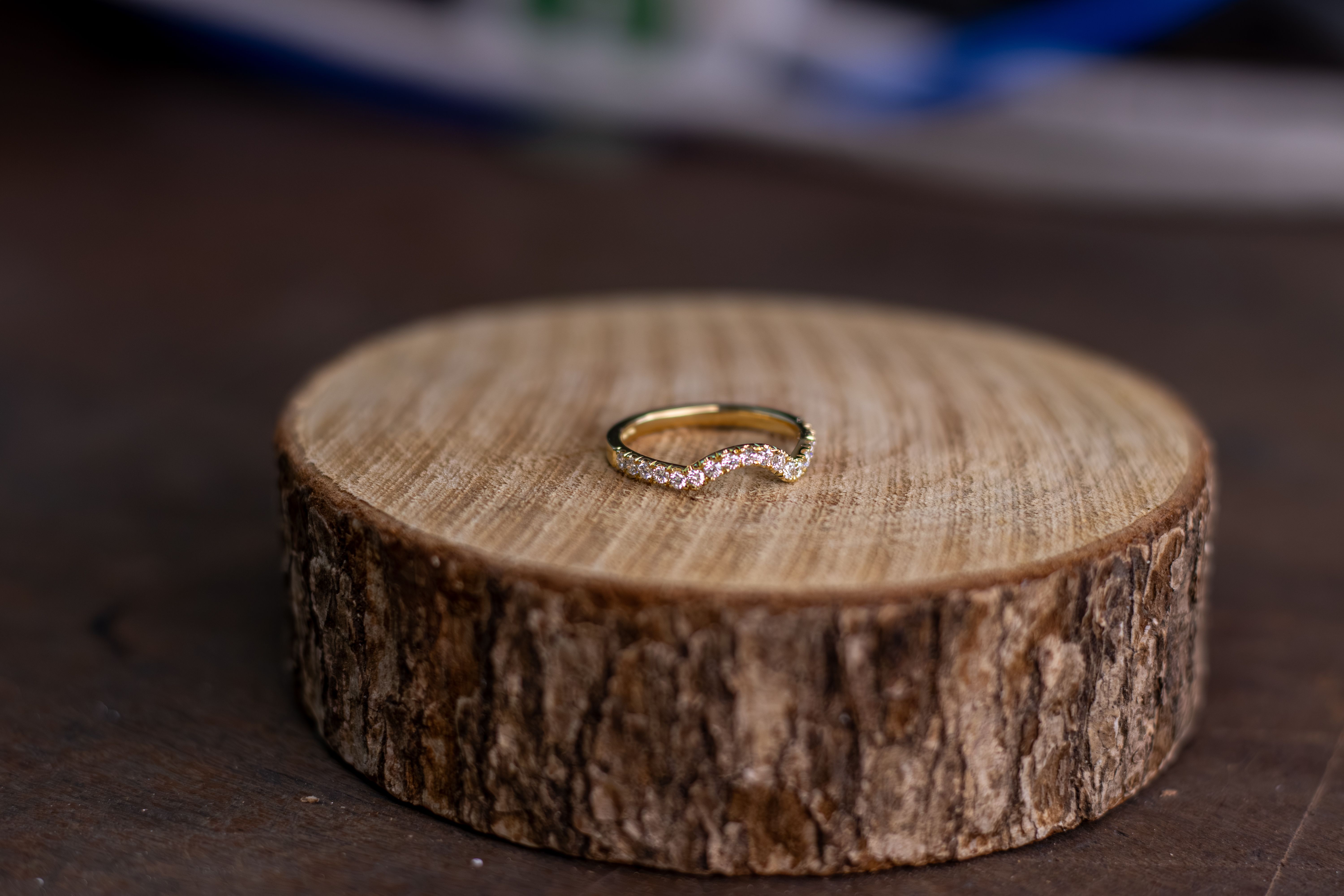 A delicate, curved gold ring adorned with small diamonds rests on a rustic wooden coaster, set against a softly blurred background.