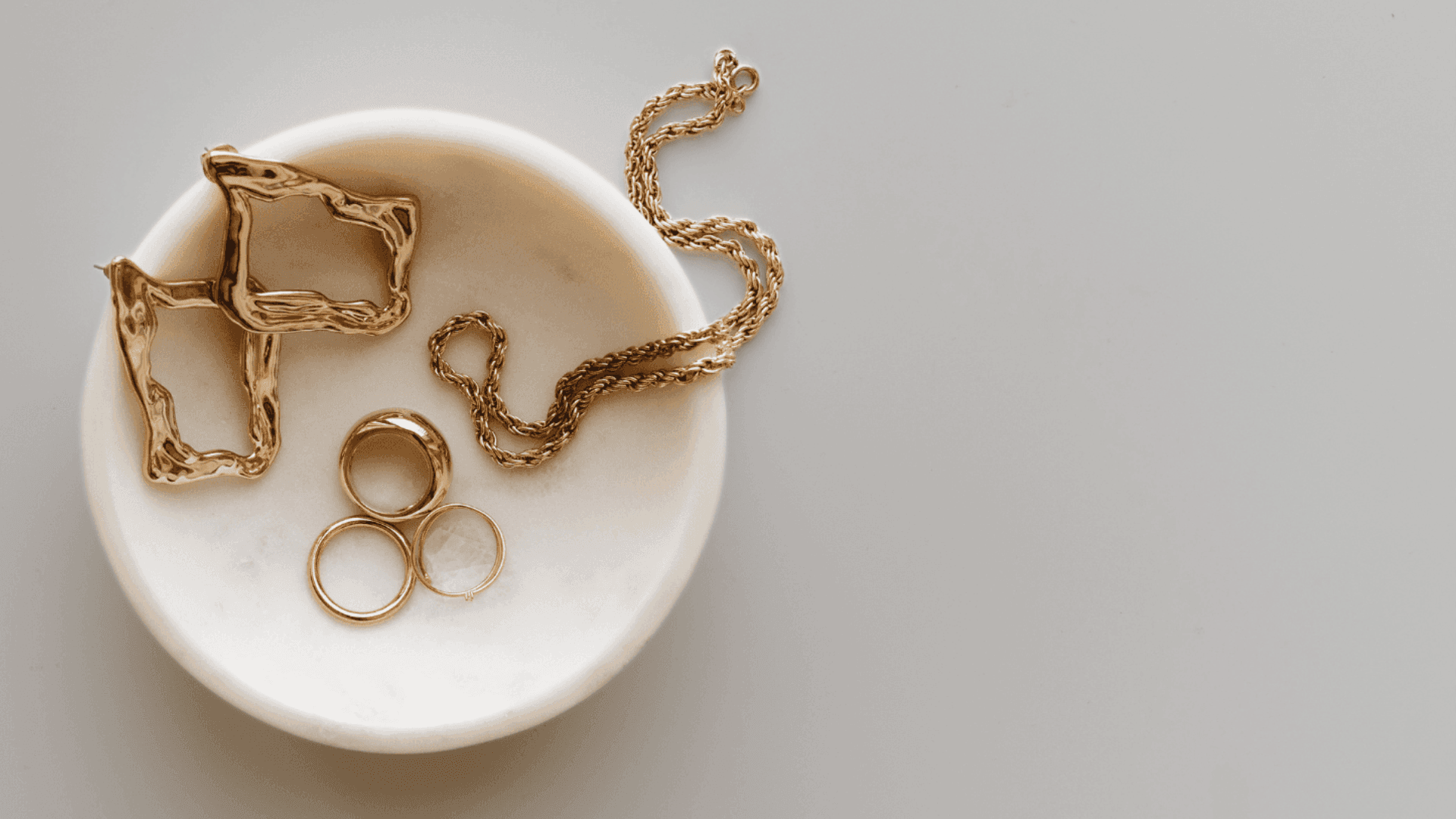 Assortment of contemporary gold jewellery, including rings, geometric earrings, and a rope chain, resting in a small, white dish on a light gray surface.