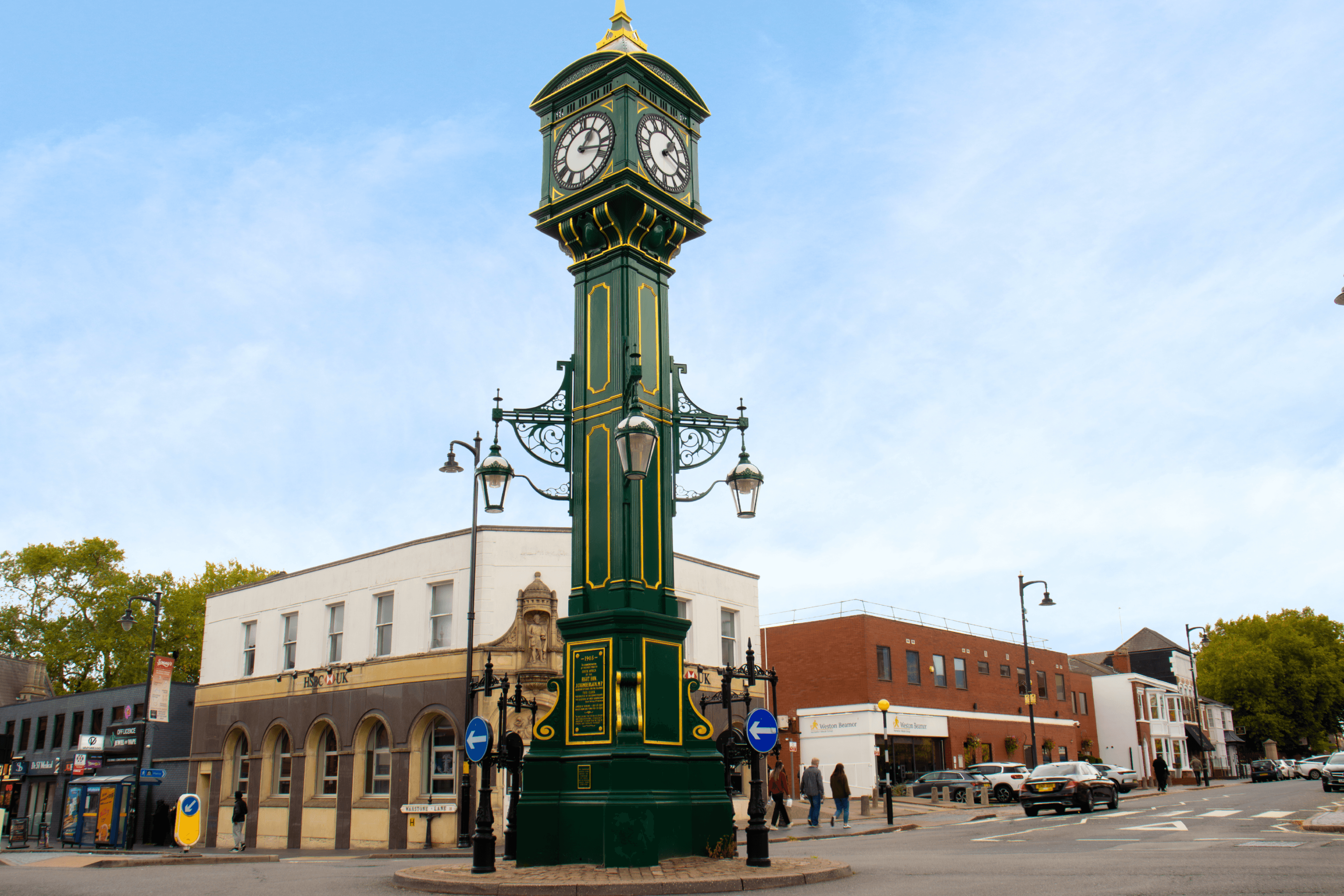 An ornate, green and gold Victorian-style clock tower stands in the middle of a street in a city.