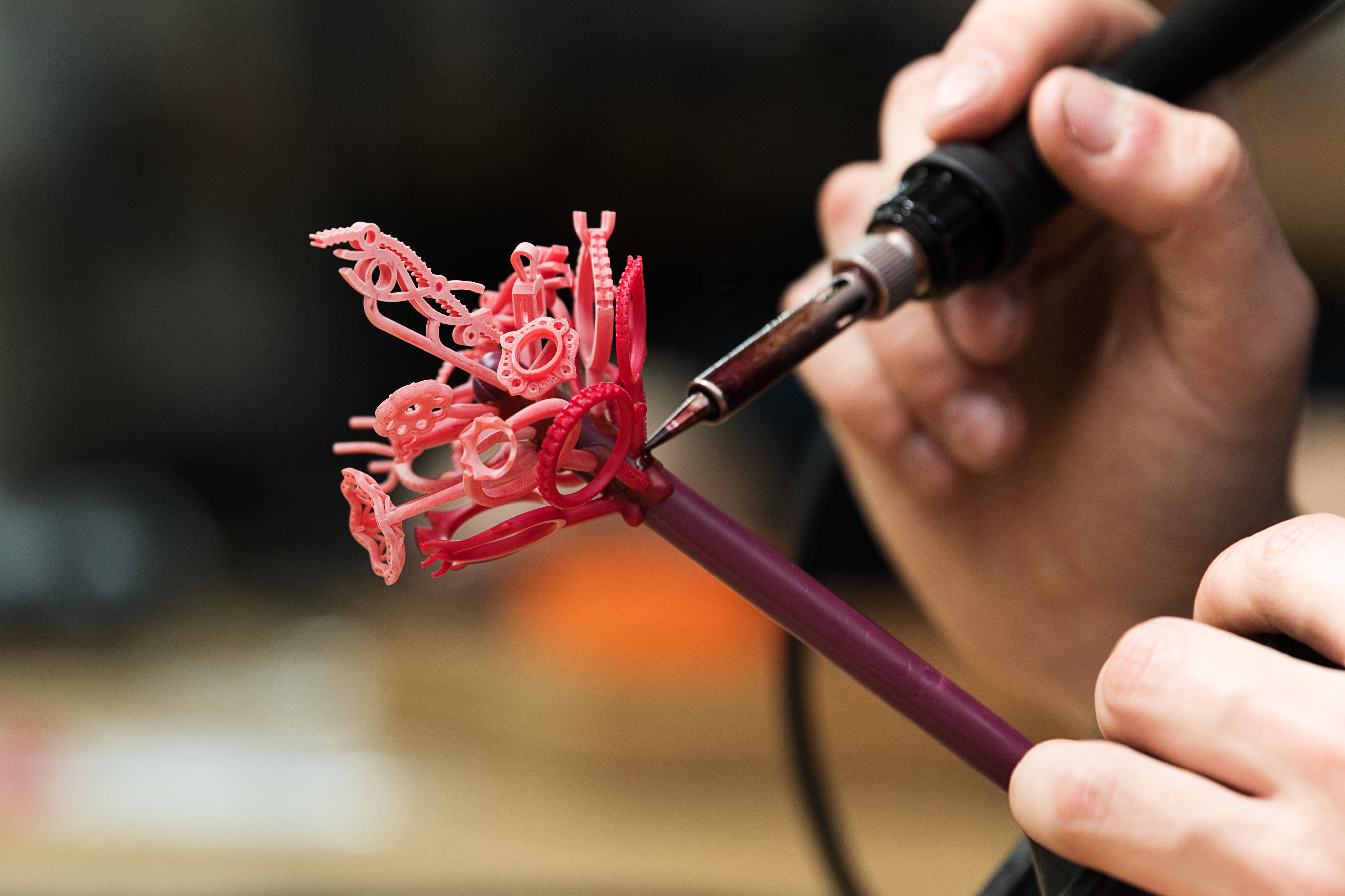 A person uses a heated tool to attach a red, complex jewelry model made of wax or resin to a larger, maroon cylindrical sprue.