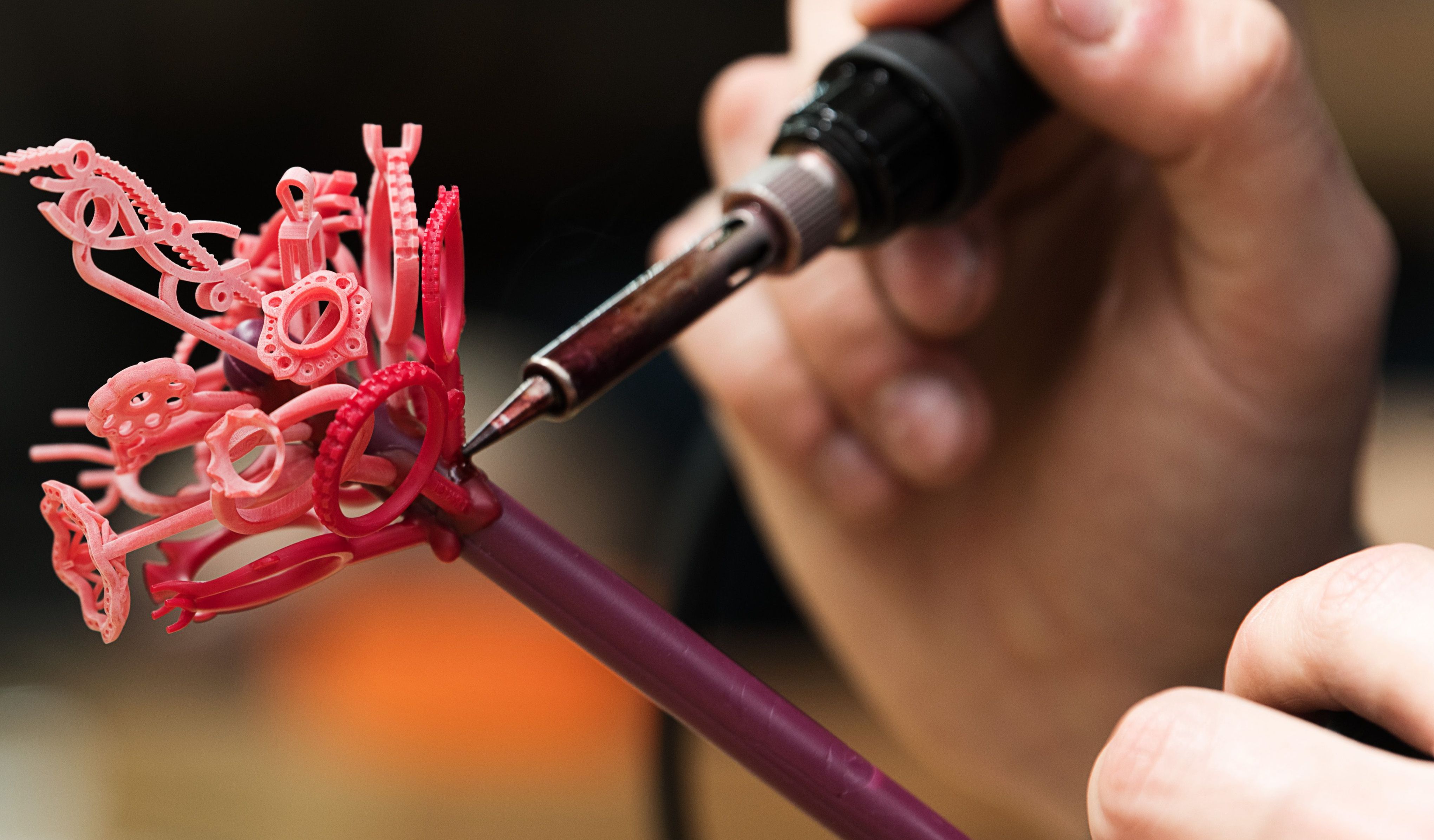 A hand uses a heated tool to attach a red wax ring to a wax "tree" covered in other jewellery models for casting.