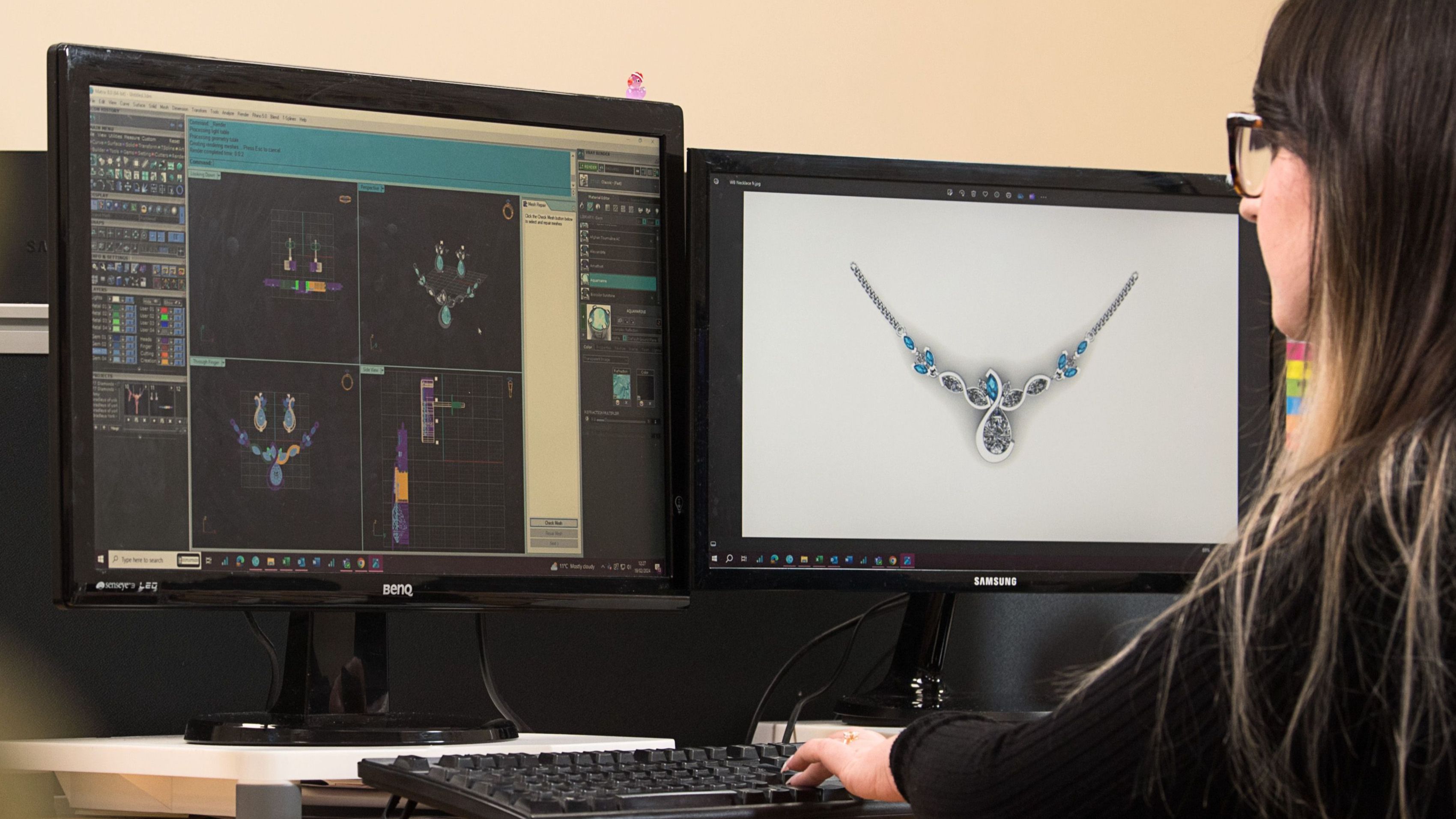 A person with dark hair and glasses is seated at a desk, looking at two computer monitors. The monitor on the right displays a 3D-rendered model of a necklace, while the monitor on the left shows a different view or design program