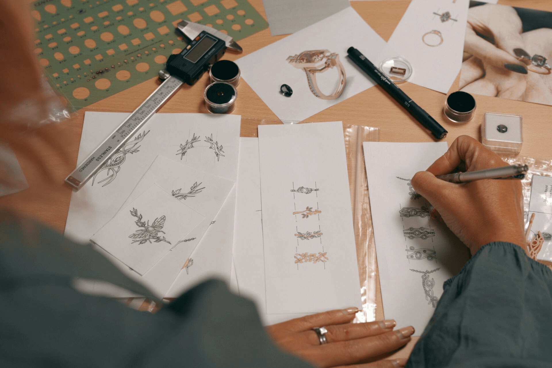 A person sketches jewelry designs at a wooden desk. Various tools are on the table, including a digital caliper, a pen, and a stencil ruler. Several sketches of rings and other jewelry are also visible on the desk.