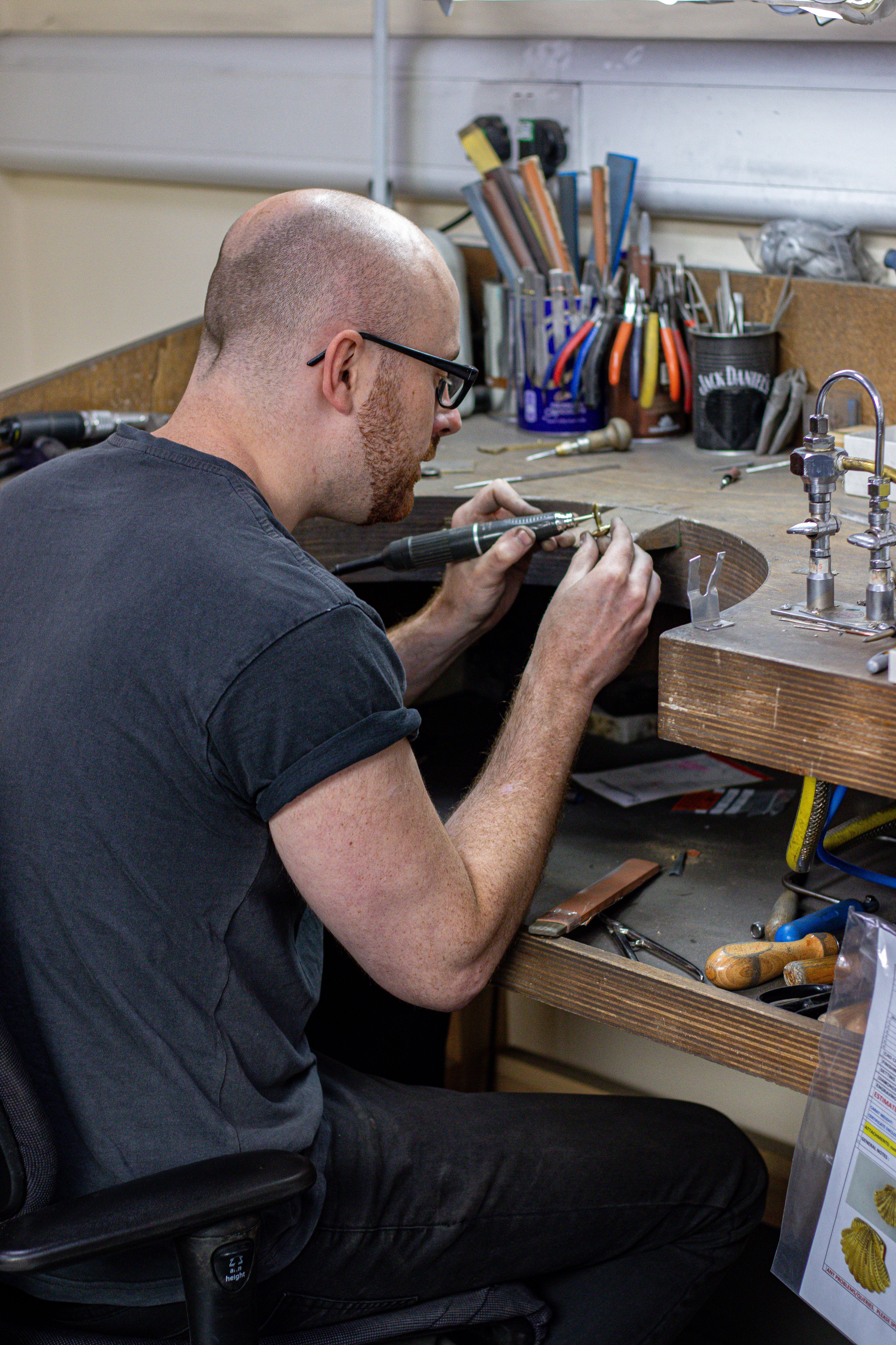 A person with glasses works intently at a cluttered workshop table, surrounded by tools. They are examining a small object under a light, focused and absorbed.
