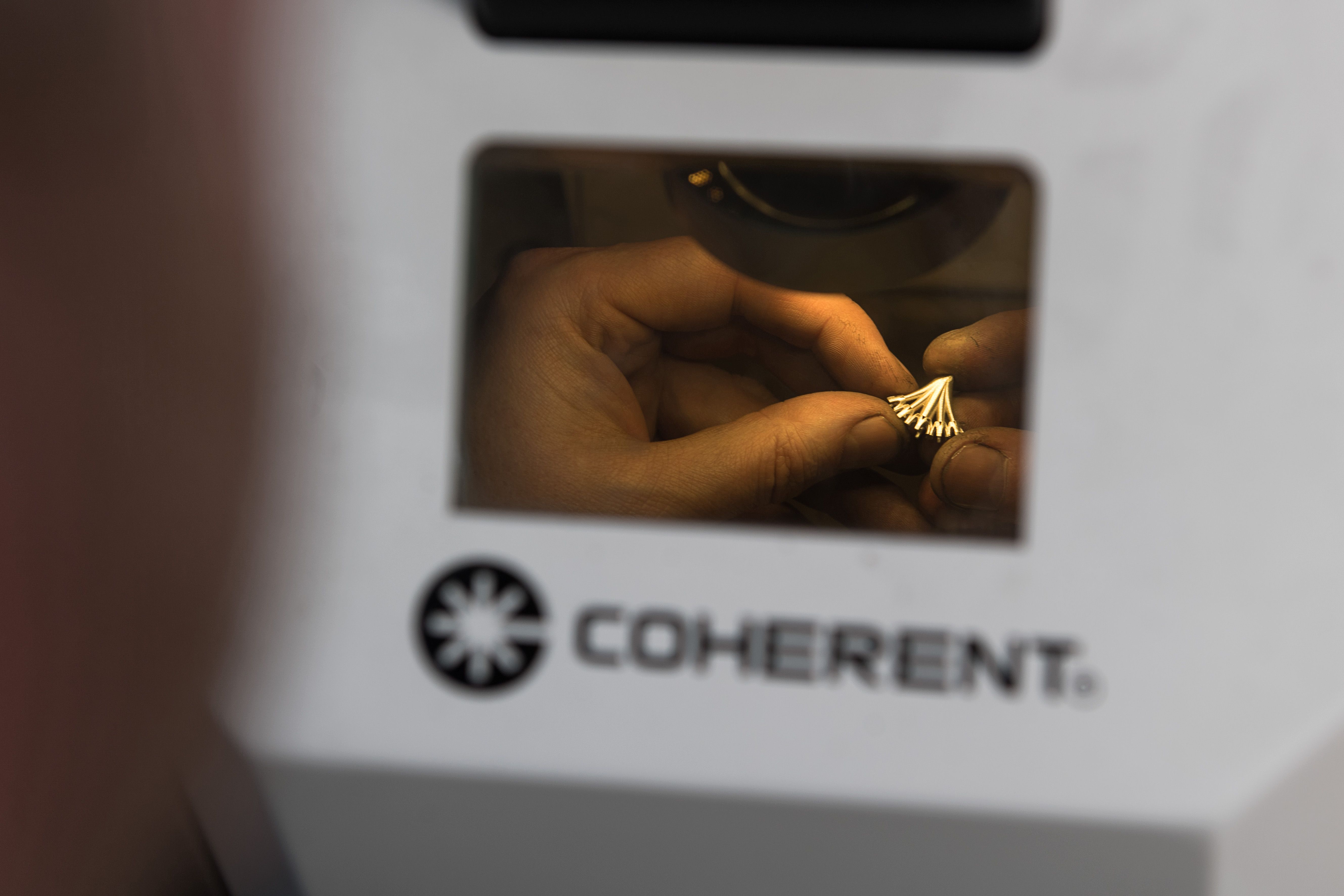 A person delicately holds a piece of jewellery beneath a Coherent laser machine, focusing intently on the precision work. The setting conveys concentration and precision.