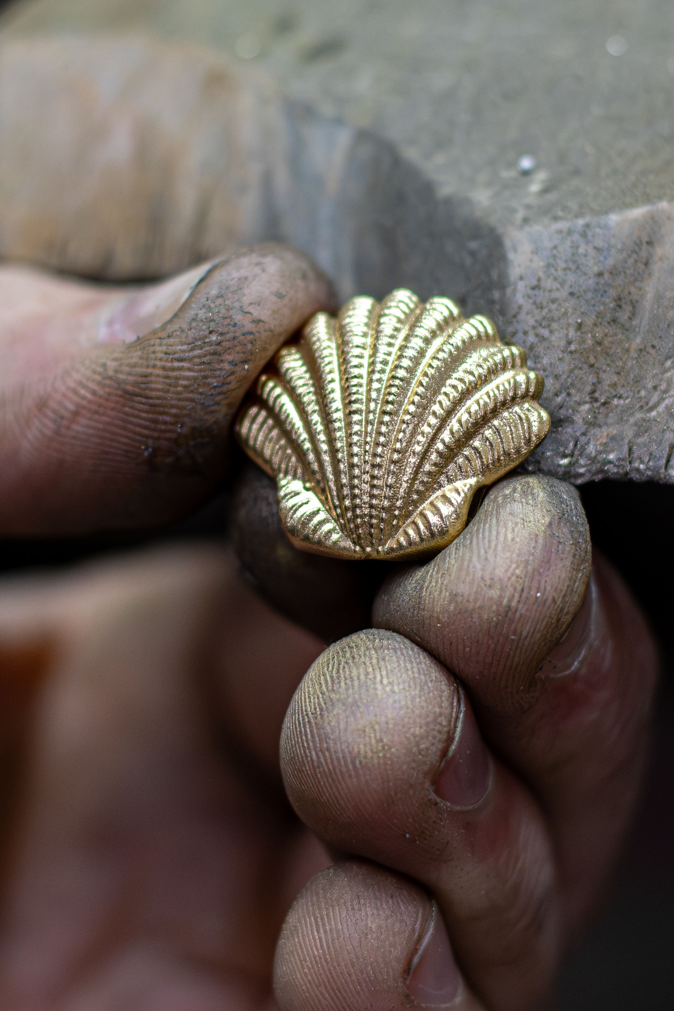 Close-up of a craftsman's hand holding a gold-colored seashell-shaped metal ornament, with visible dust and texture from the workbench.
