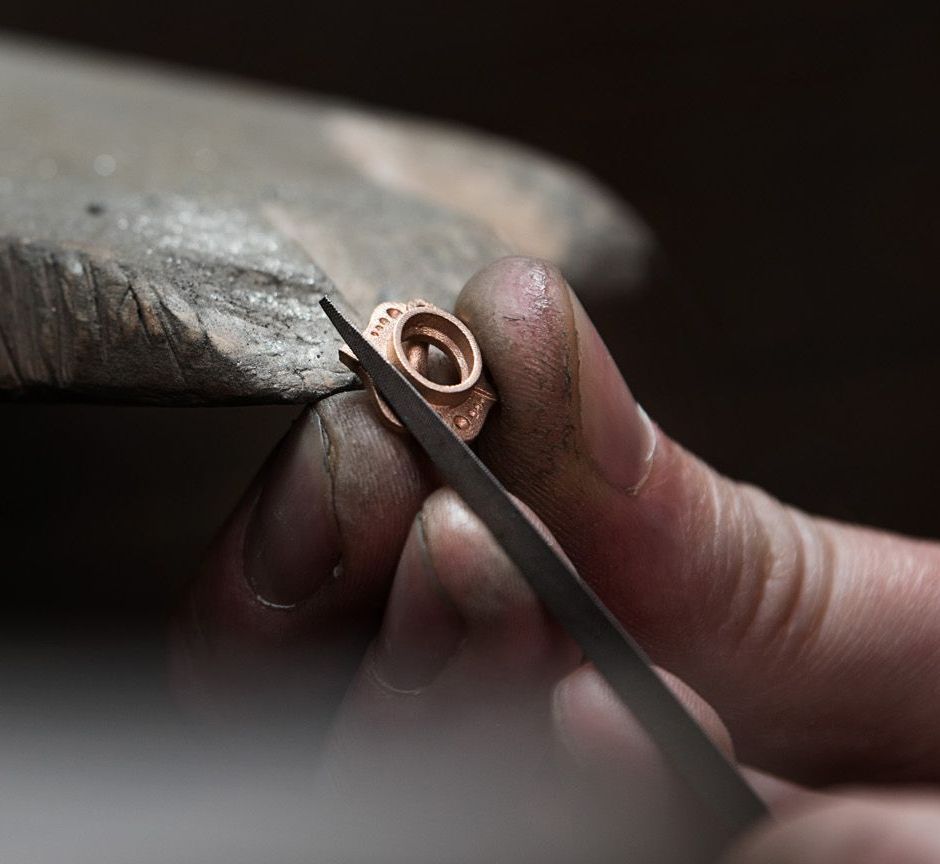 A jeweller’s hands are seen holding a small, intricate metal piece on a workbench, carefully filing it by hand with a small tool.