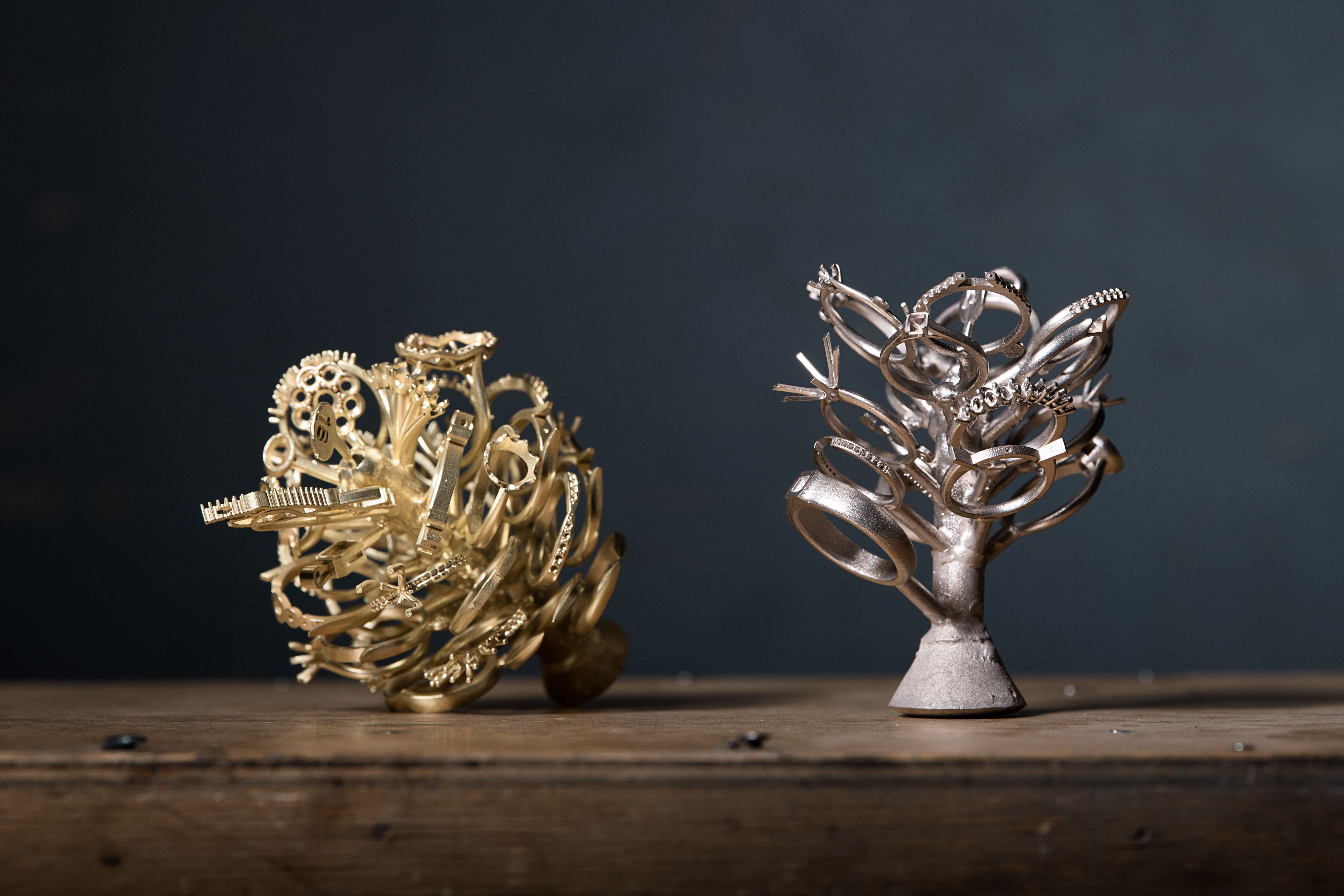 Two intricate metal sculptures rest on a wooden surface against a dark background. The left piece is gold-toned and spherical, while the right is silver-toned and tree-like.