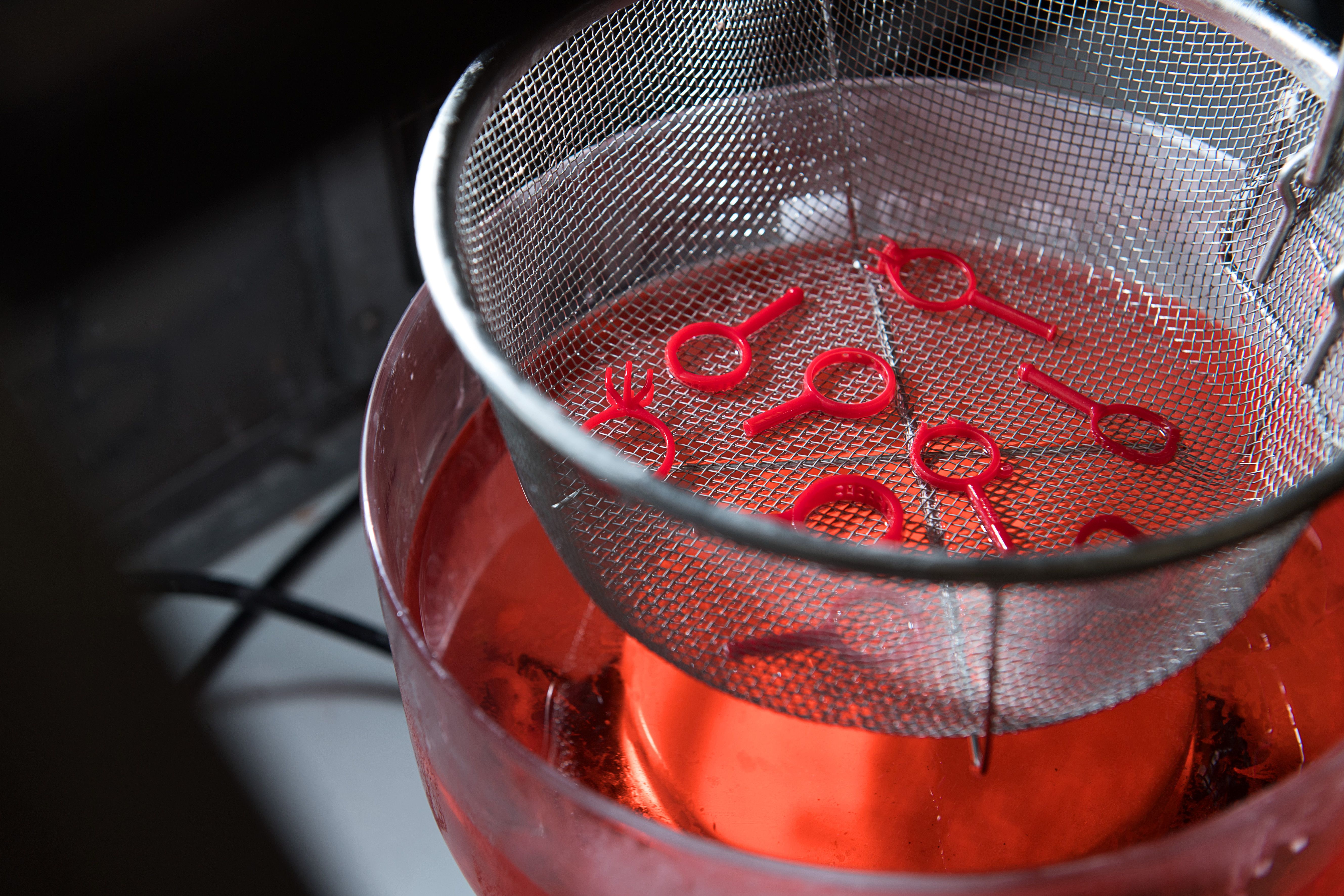 A metal strainer with red plastic ring shapes is submerged in a container filled with a red liquid, conveying an experimental or industrial setting.
