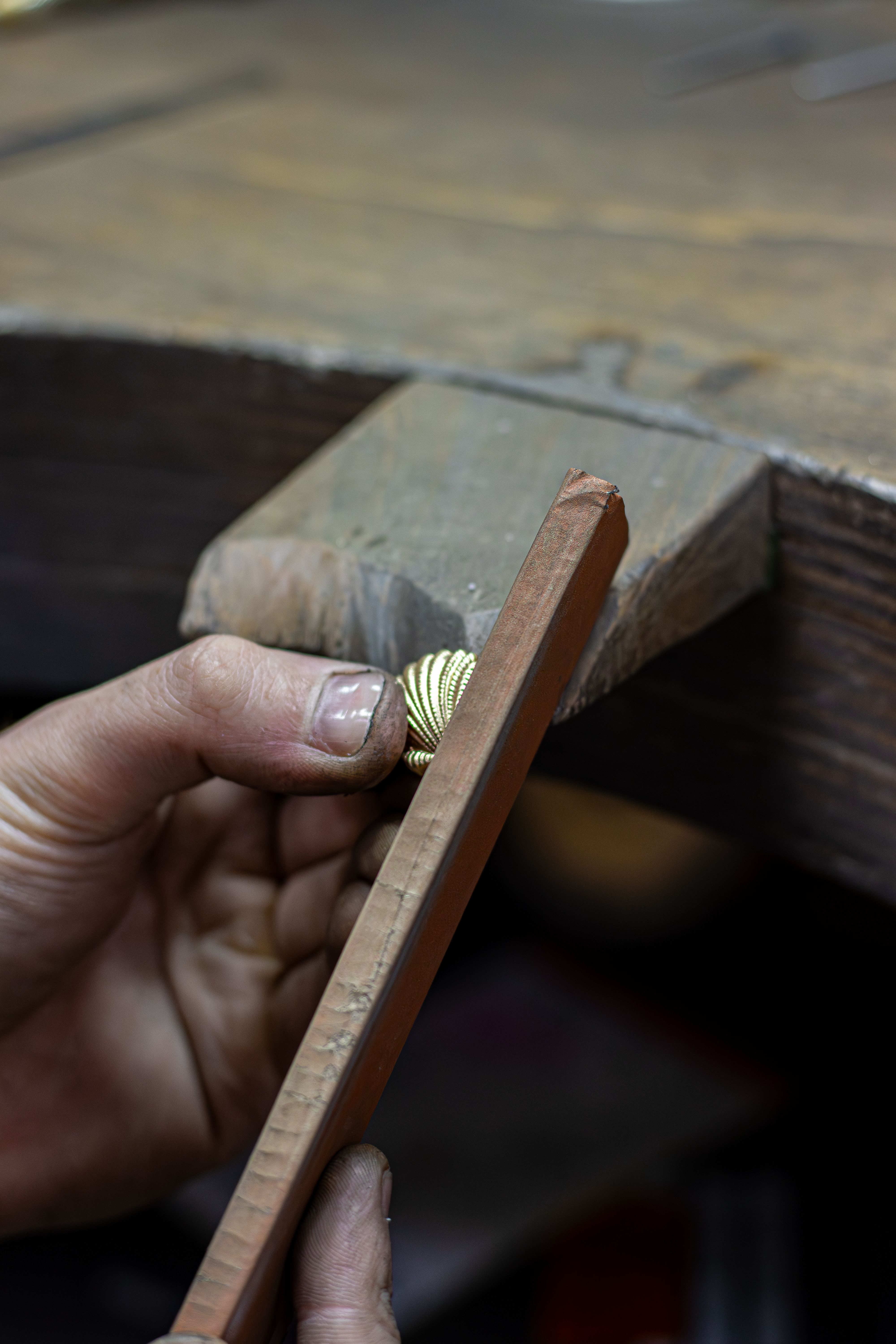 A person uses a file to shape or smooth a small object held in their hand on a wooden workbench.