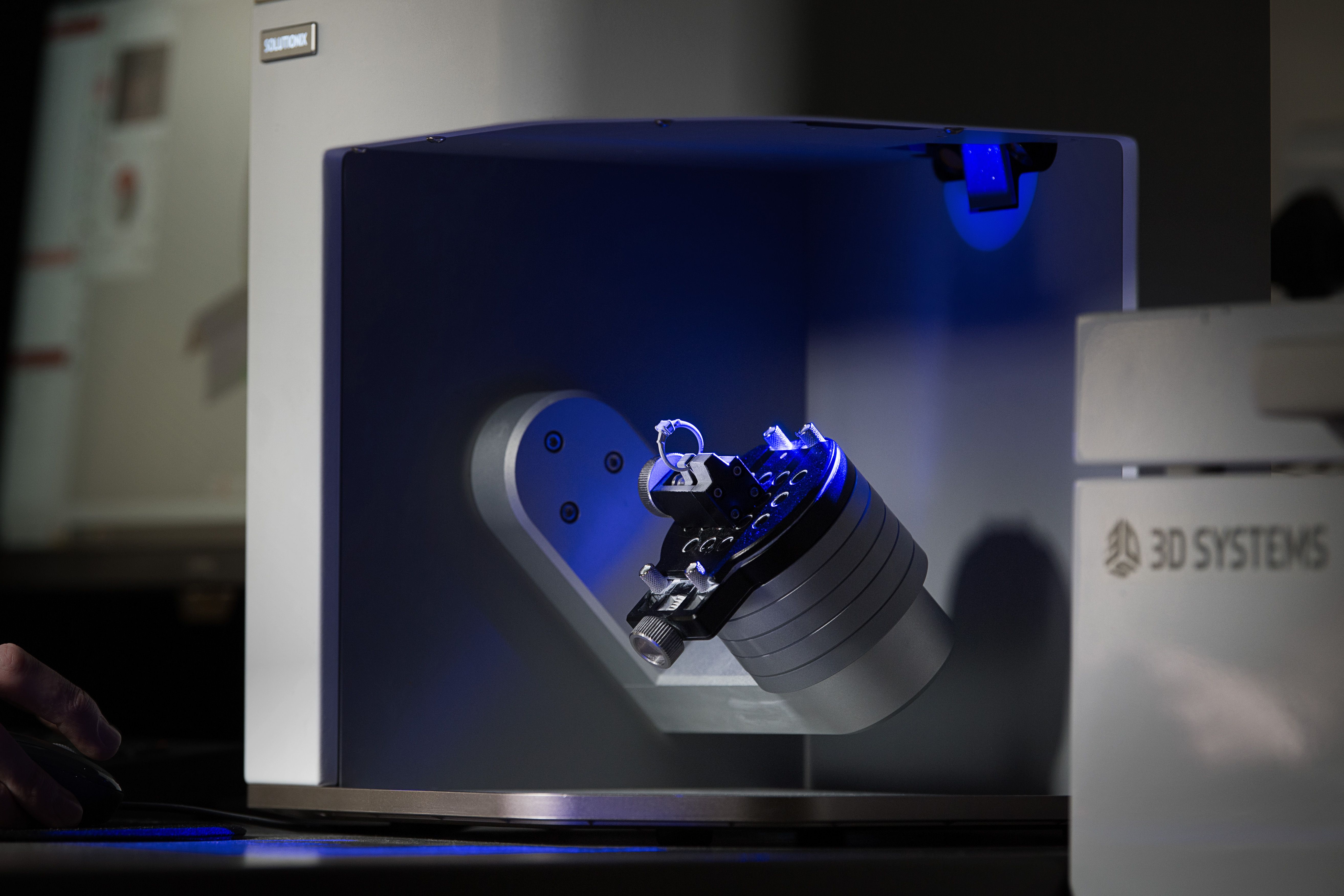 A small object is being scanned by a 3D scanner with a blue light. The object is a small model attached to a cylindrical base. The scanner is a white machine with "3D SYSTEMS" visible on the front.