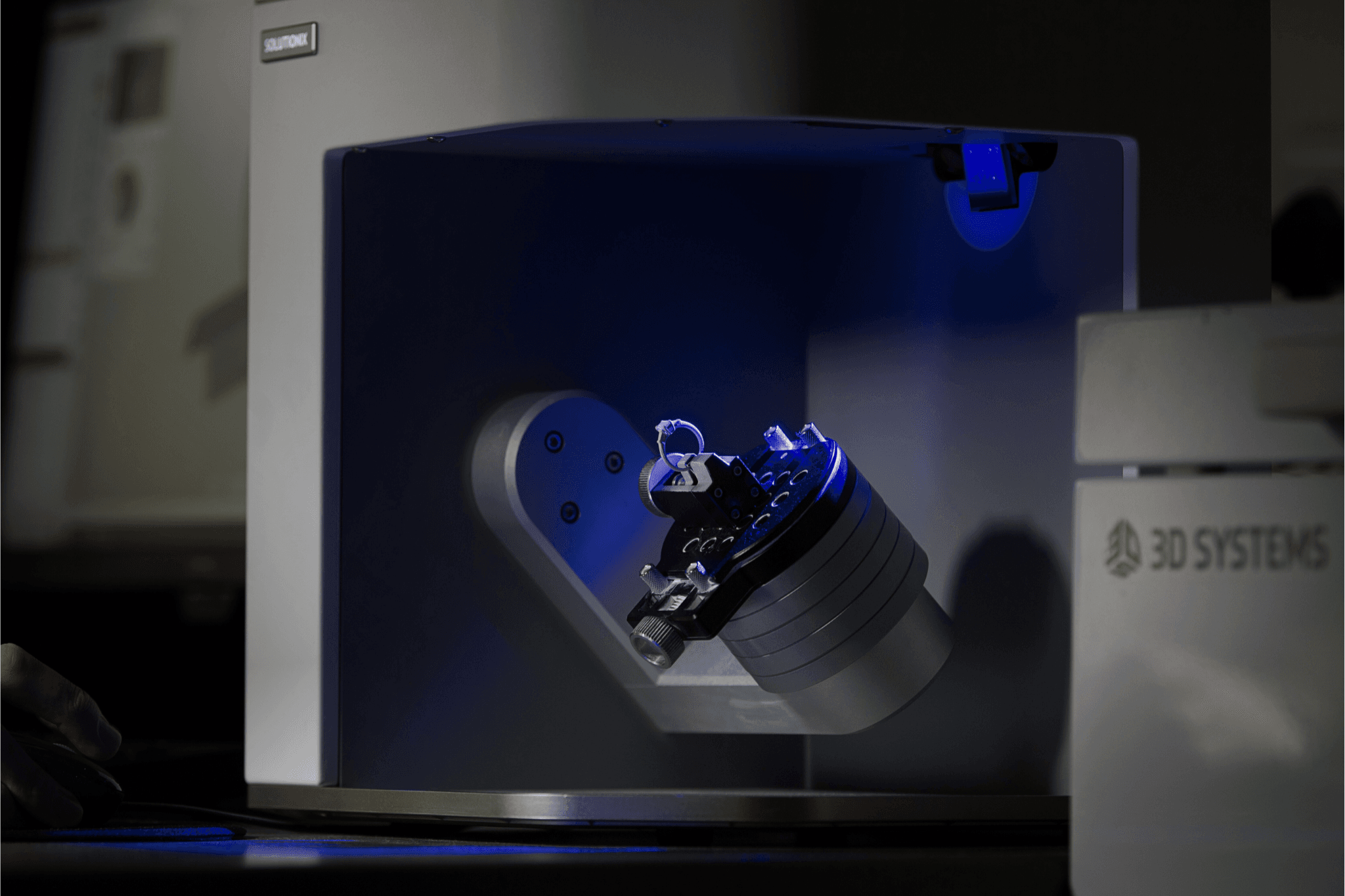 A dark, eye-level shot shows a 3D scanner with a blue light scanning a small, intricate model. The model is attached to a cylindrical base that is tilted at an angle.