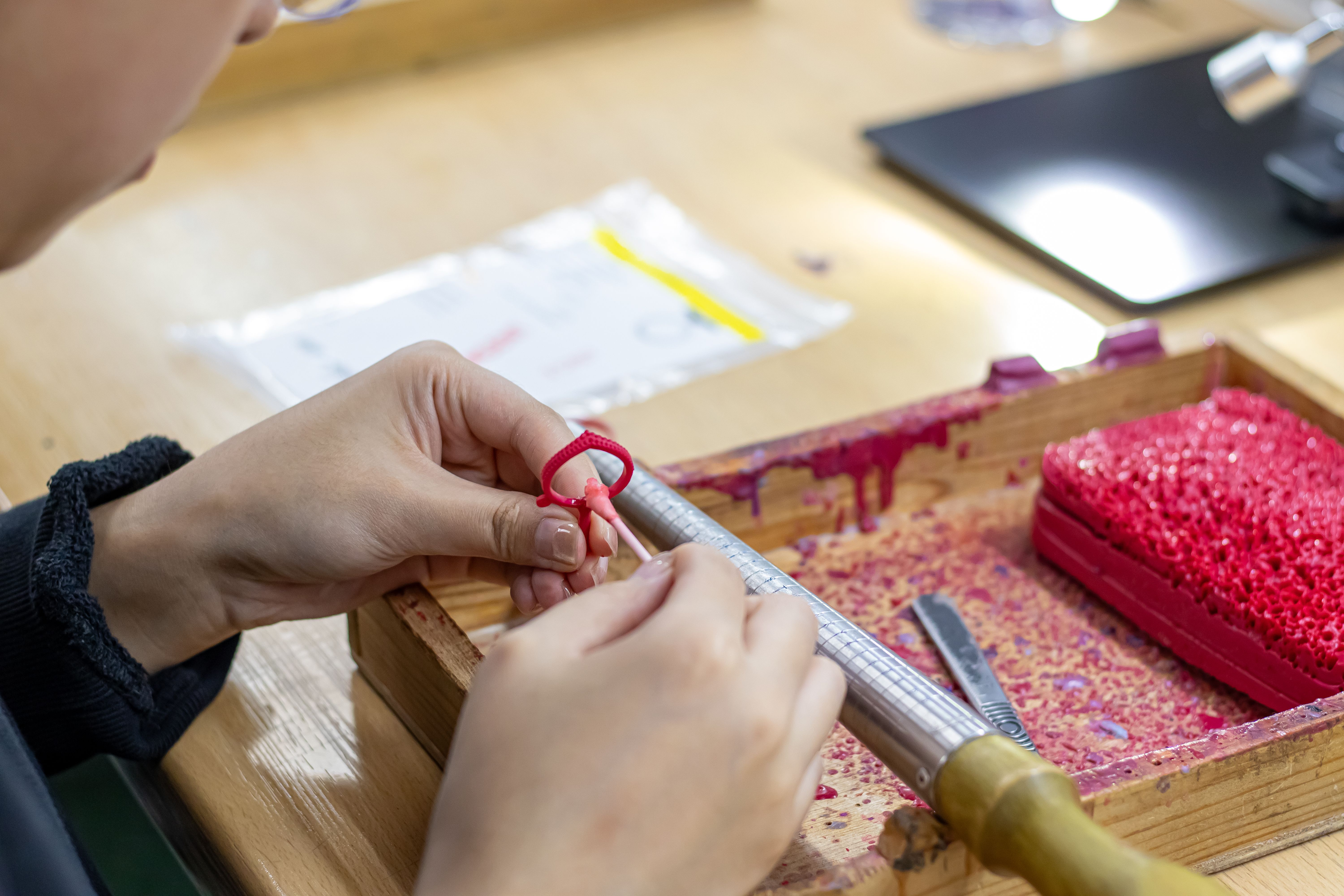 A close-up of a person's hands using a small tool to work on a red wax model of a ring at a workbench.