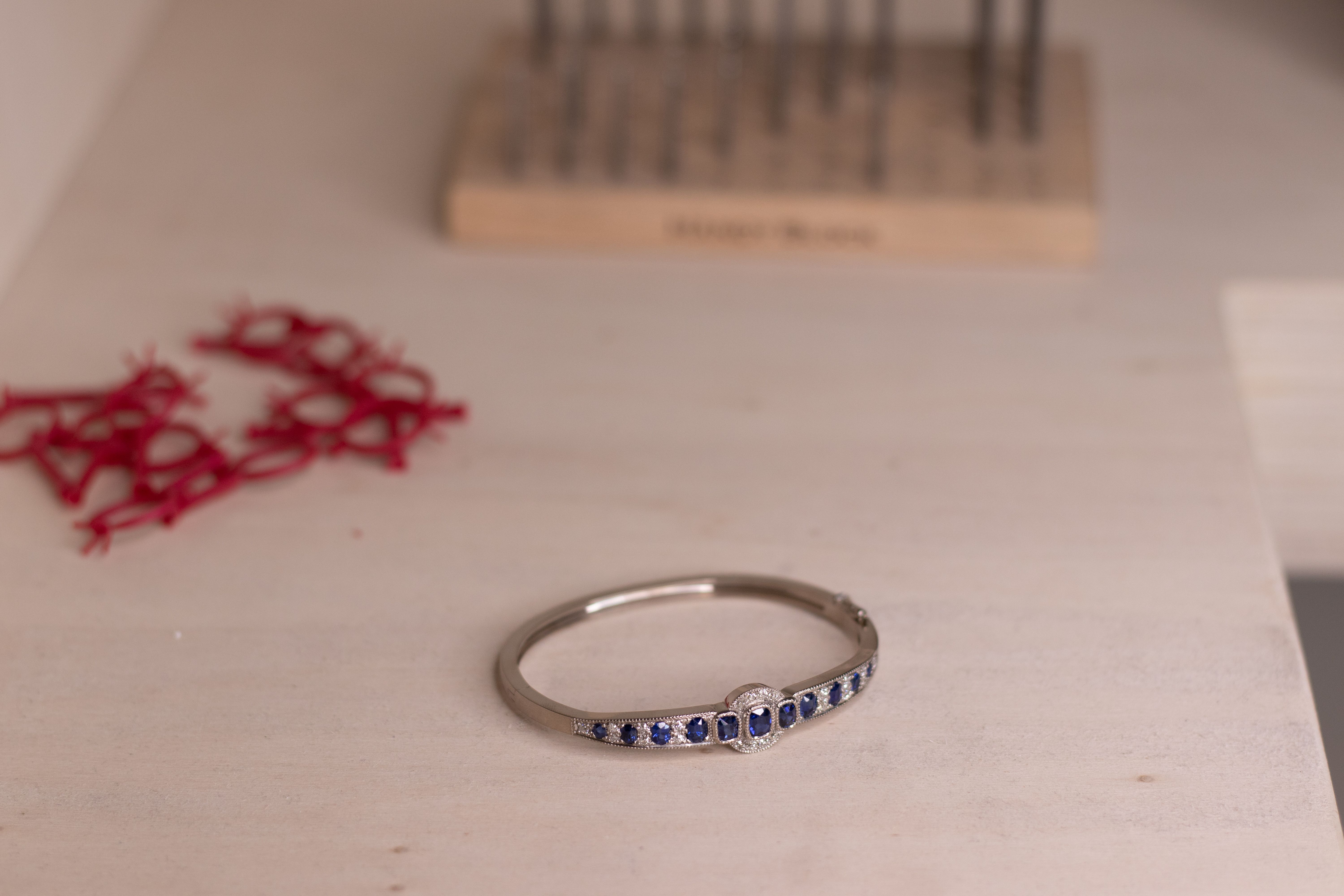 A luxury sapphire and diamond hinged bangle featuring a central oval sapphire cluster, shown in a jewellery workshop setting.