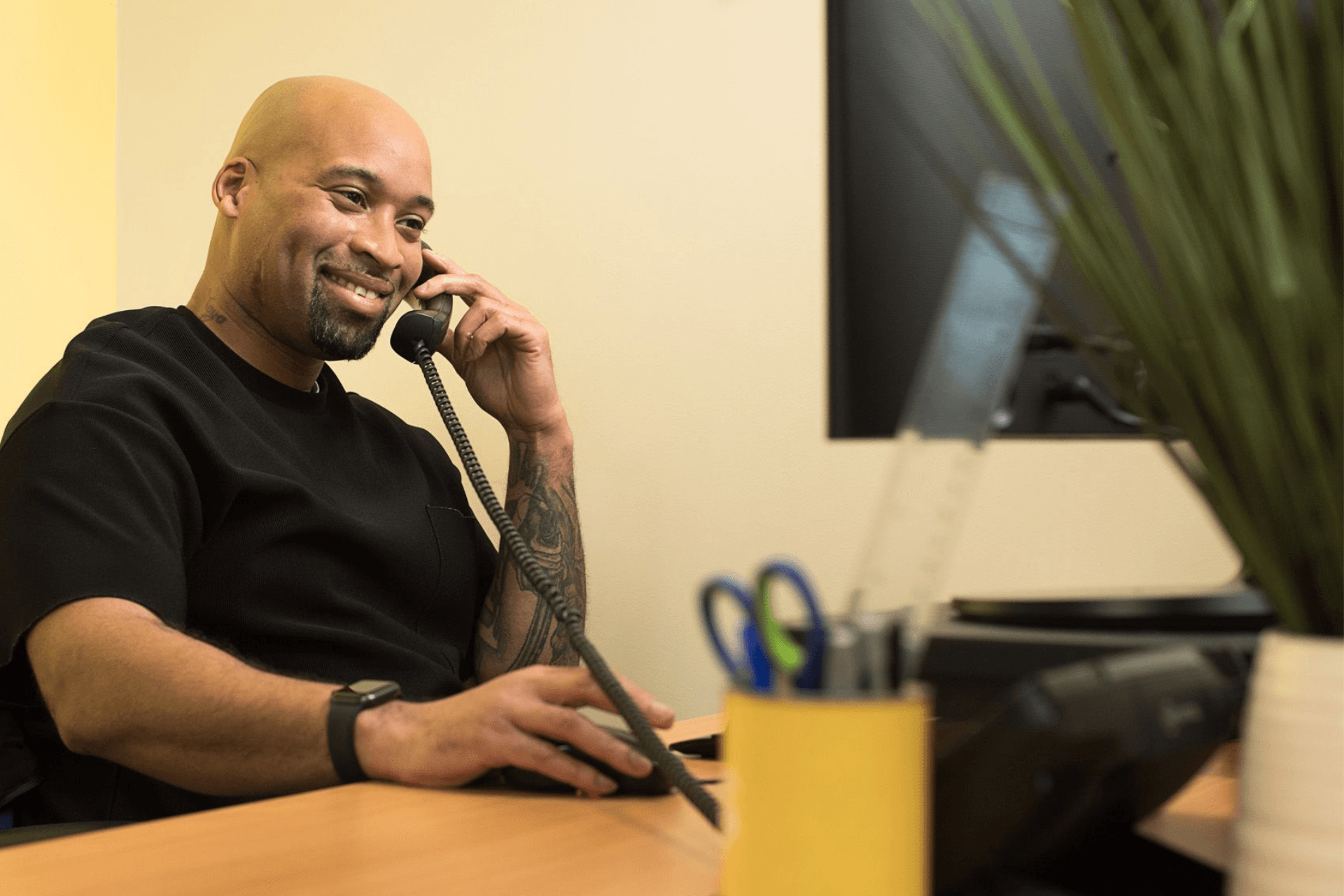A smiling man with tattoos on his arm sits at a wooden desk in an office, holding a landline phone to his ear. He is wearing a black shirt and a watch, and in the background, there's a computer monitor and a pot of pens.