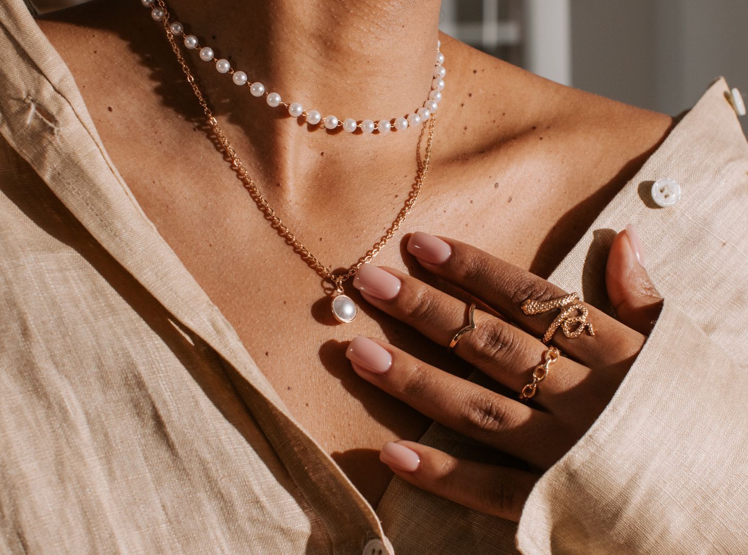 A woman wearing a beige shirt with pearl and gold necklaces, adorned with elegant rings on pink-manicured fingers. The mood is chic and sophisticated.