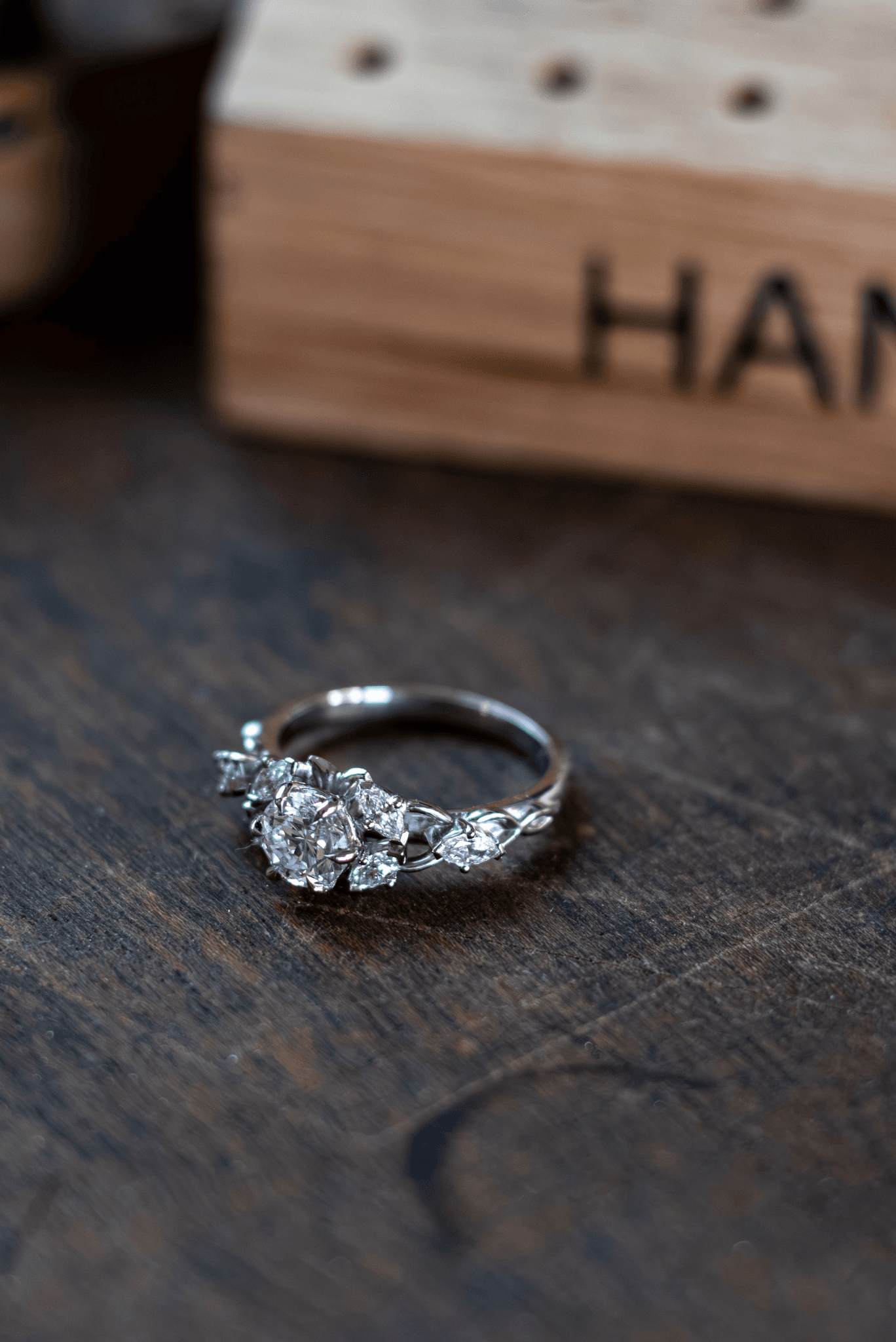 A close-up, horizontal shot of a silver or platinum ring with a large central diamond and smaller accent diamonds on a textured wooden surface.