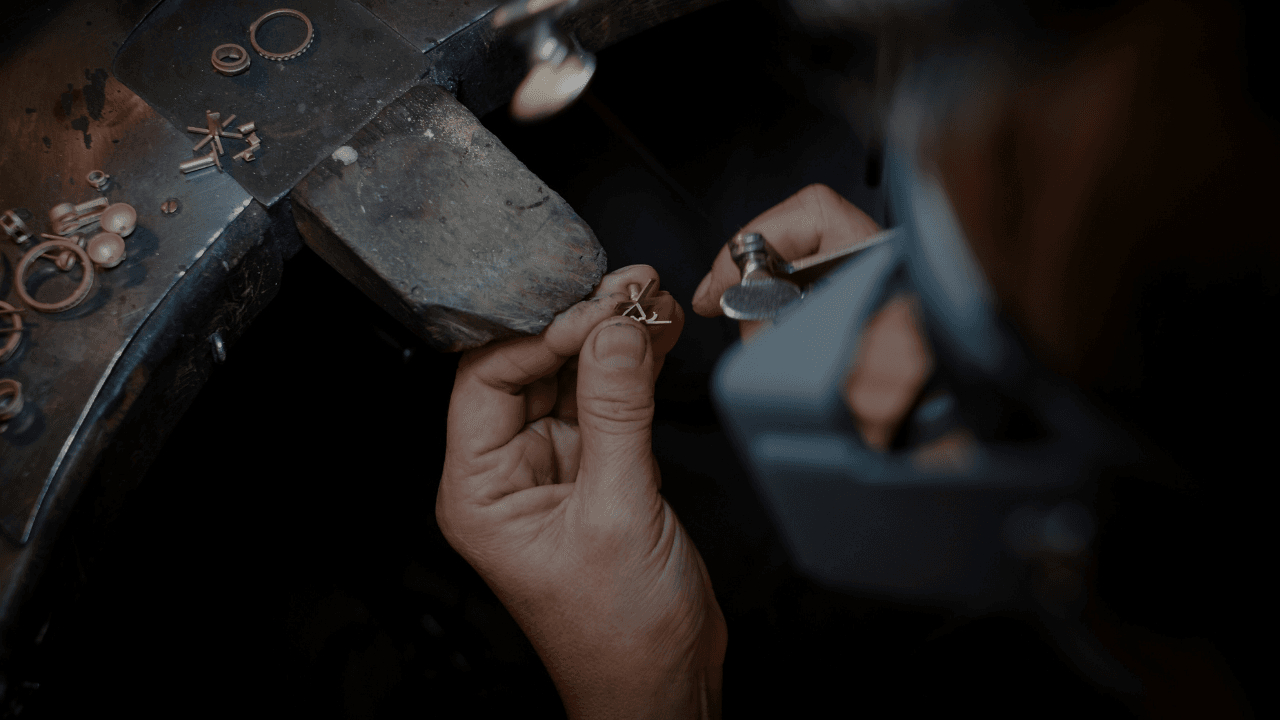 A close-up shows a person's hands meticulously working on a small piece of jewelry with tools, resting on a wooden jeweler's bench peg. Other small metal components are scattered nearby.