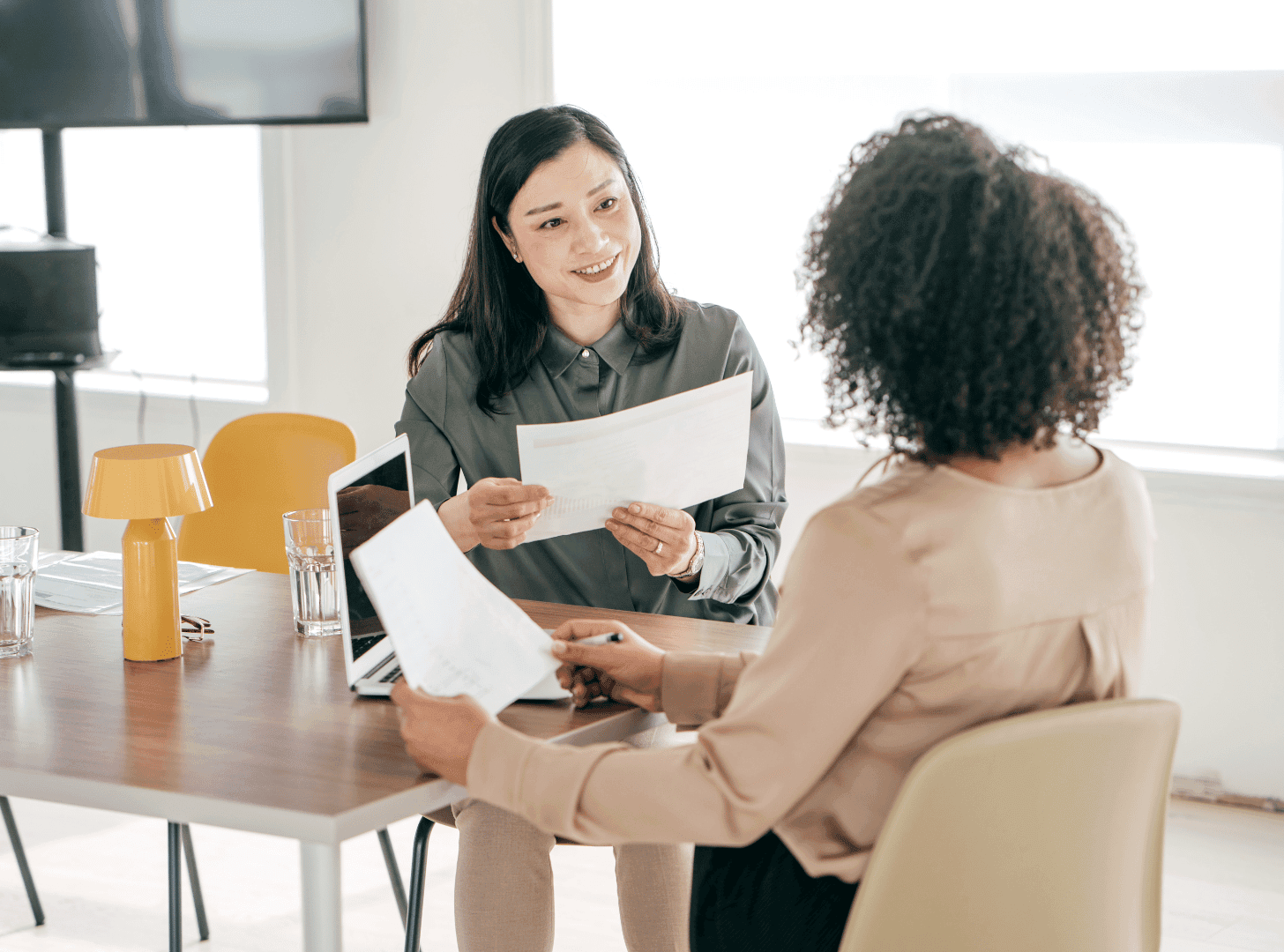 Two professional women in a brightly lit office sitting at a wooden desk, discussing jewellery design specifications and reviewing documents during a client consultation.