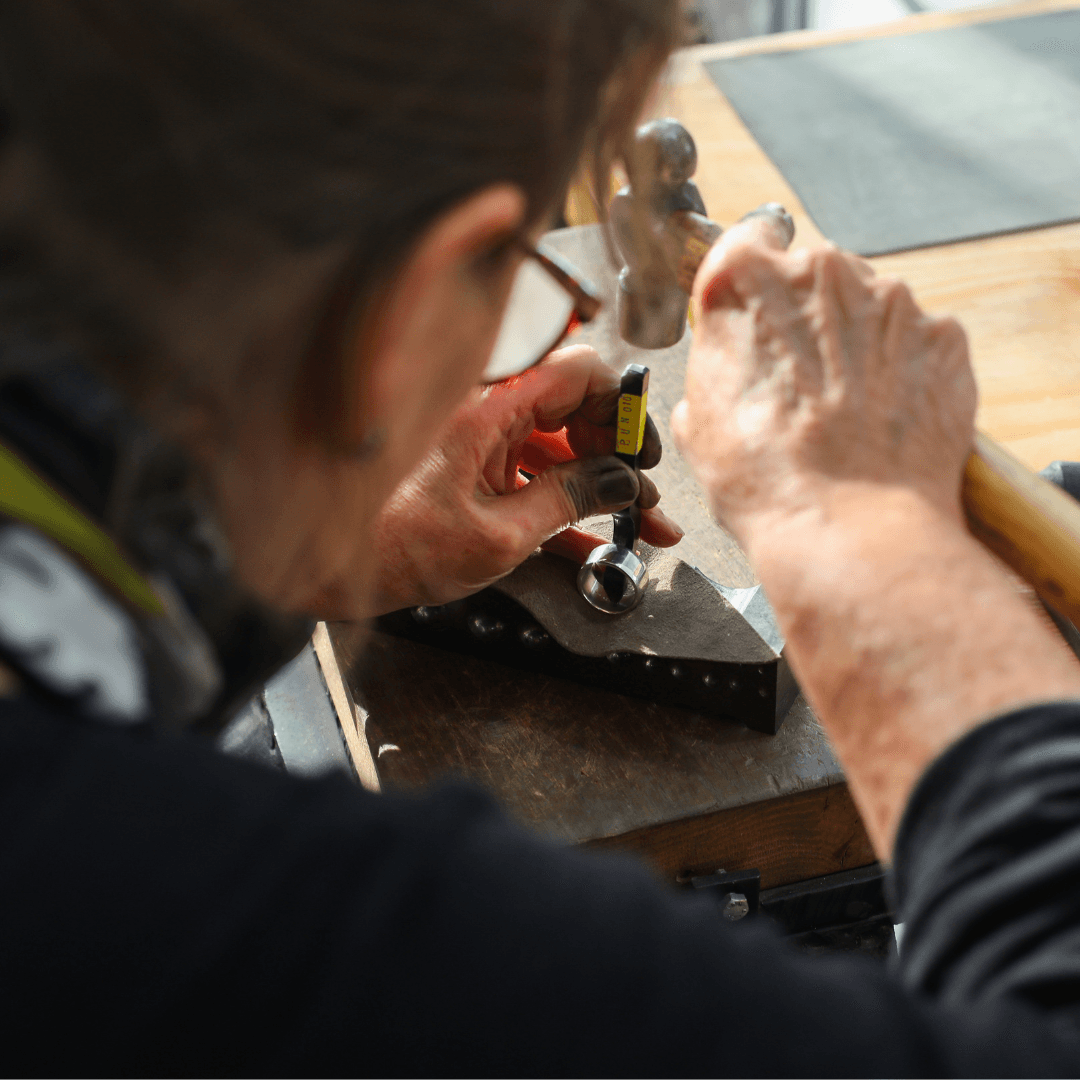 A close-up, over-the-shoulder shot shows a person wearing glasses and a black shirt as they use a hammer to strike a hallmark punch. The punch is held over a metal signet ring, which is resting on a steel block on a wooden workbench.