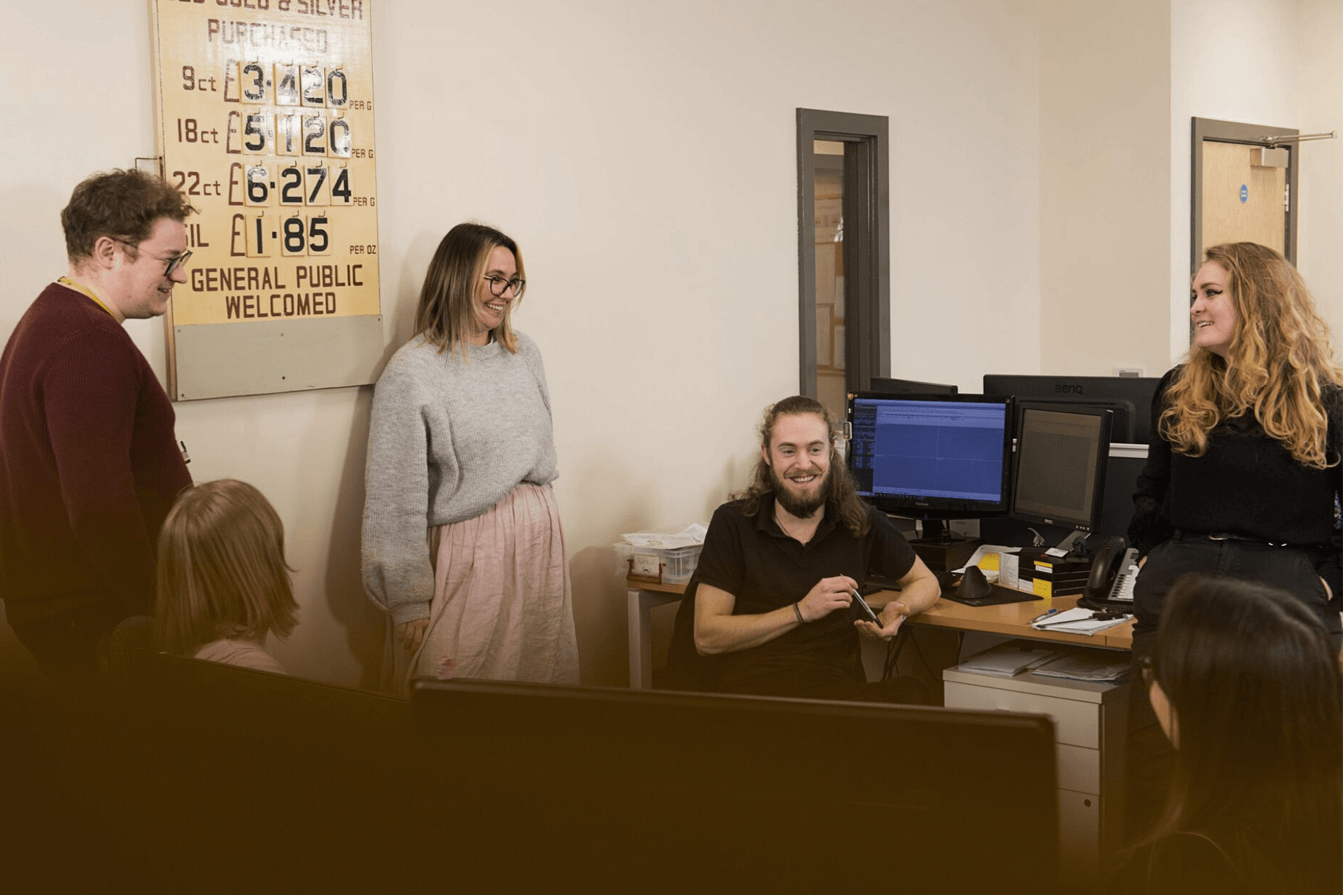 Four adults and a young child are gathered in an office. Three of the adults are standing near a wall with a sign displaying weights and prices, while a fourth adult is sitting at a desk with dual monitors.