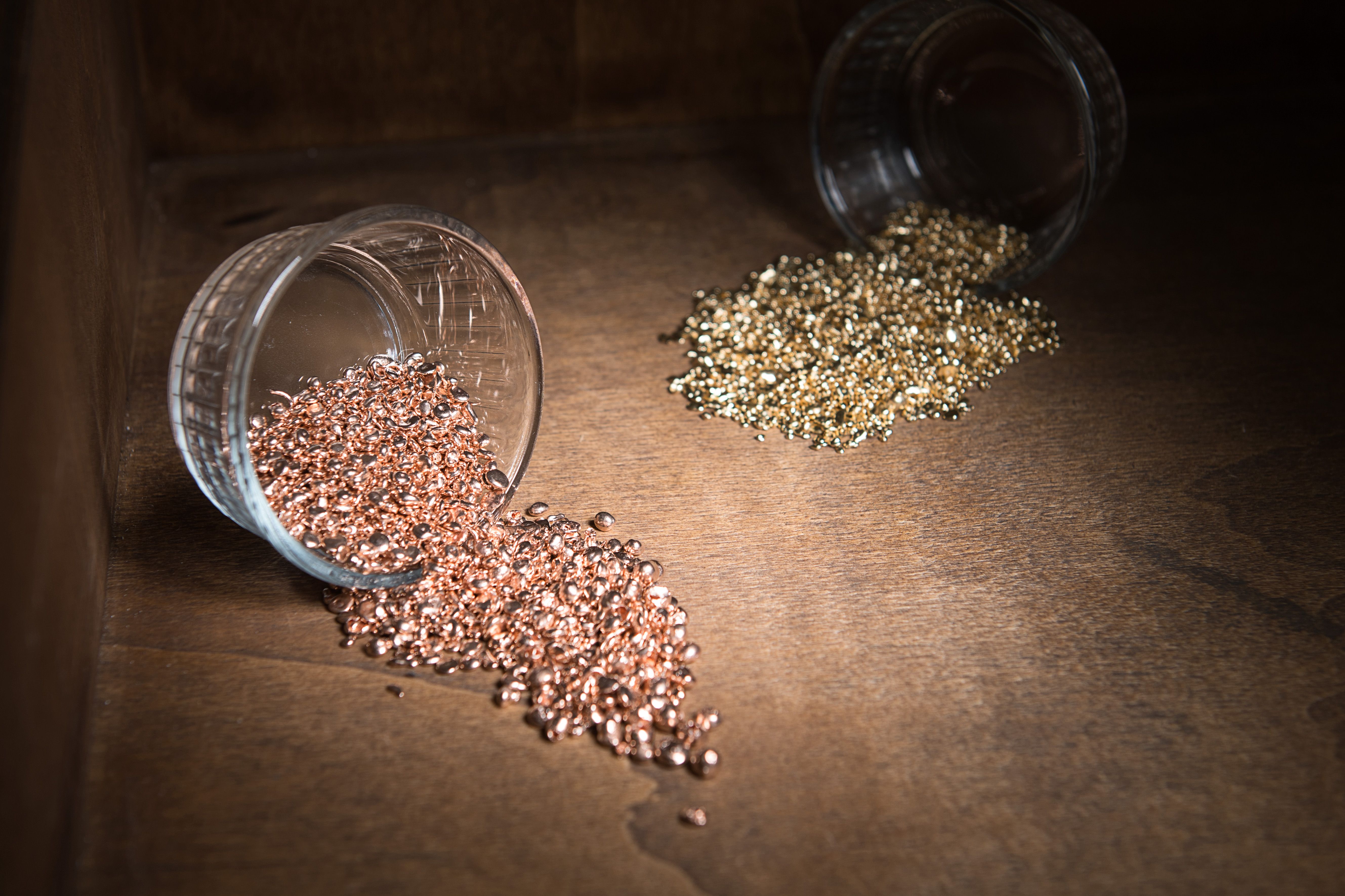 Two glass containers lie on their sides on a wooden surface, with small, shiny metal pellets spilling out. One container holds gold-colored pellets, while the other holds rose gold or copper-colored pellets.