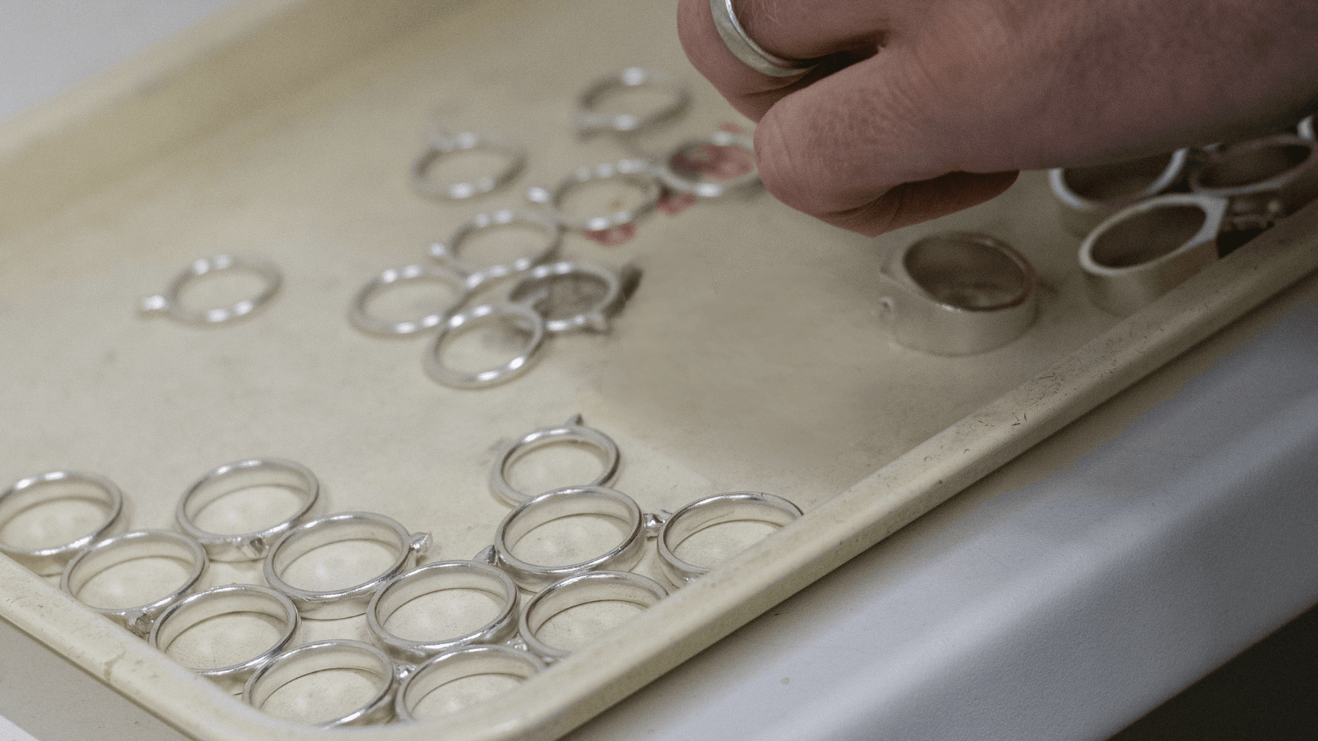 Jewellery technician's hand inspecting a tray of unfinished silver or white gold ring castings in a manufacturing facility.
